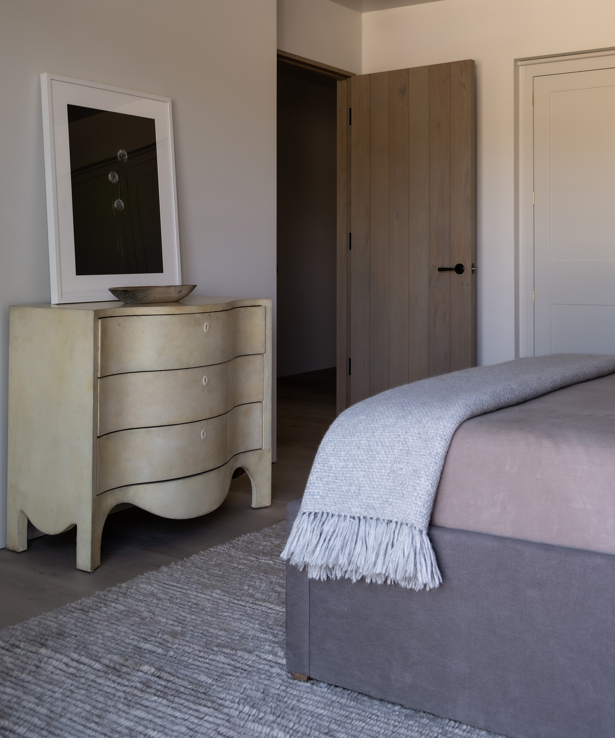 A neatly made bed with a gray blanket sits beside a light-colored dresser topped with a bowl and framed mirror, creating a serene setting in this minimalist bedroom remodel featuring neutral colors and an open wooden door.