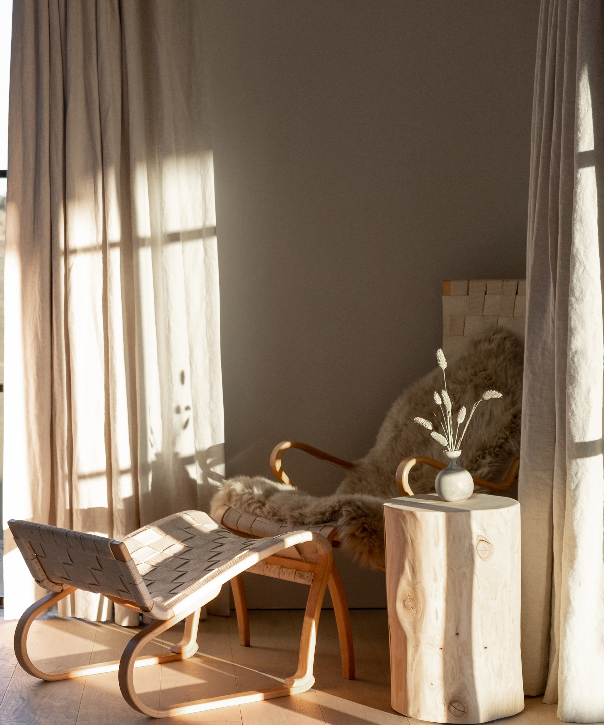 A cozy corner with a wooden lounge chair draped in a fur throw, a matching ottoman, and a stump side table holding a small vase with dried flowers—sunlit ambiance inspired by Galerie Provenance elegance.