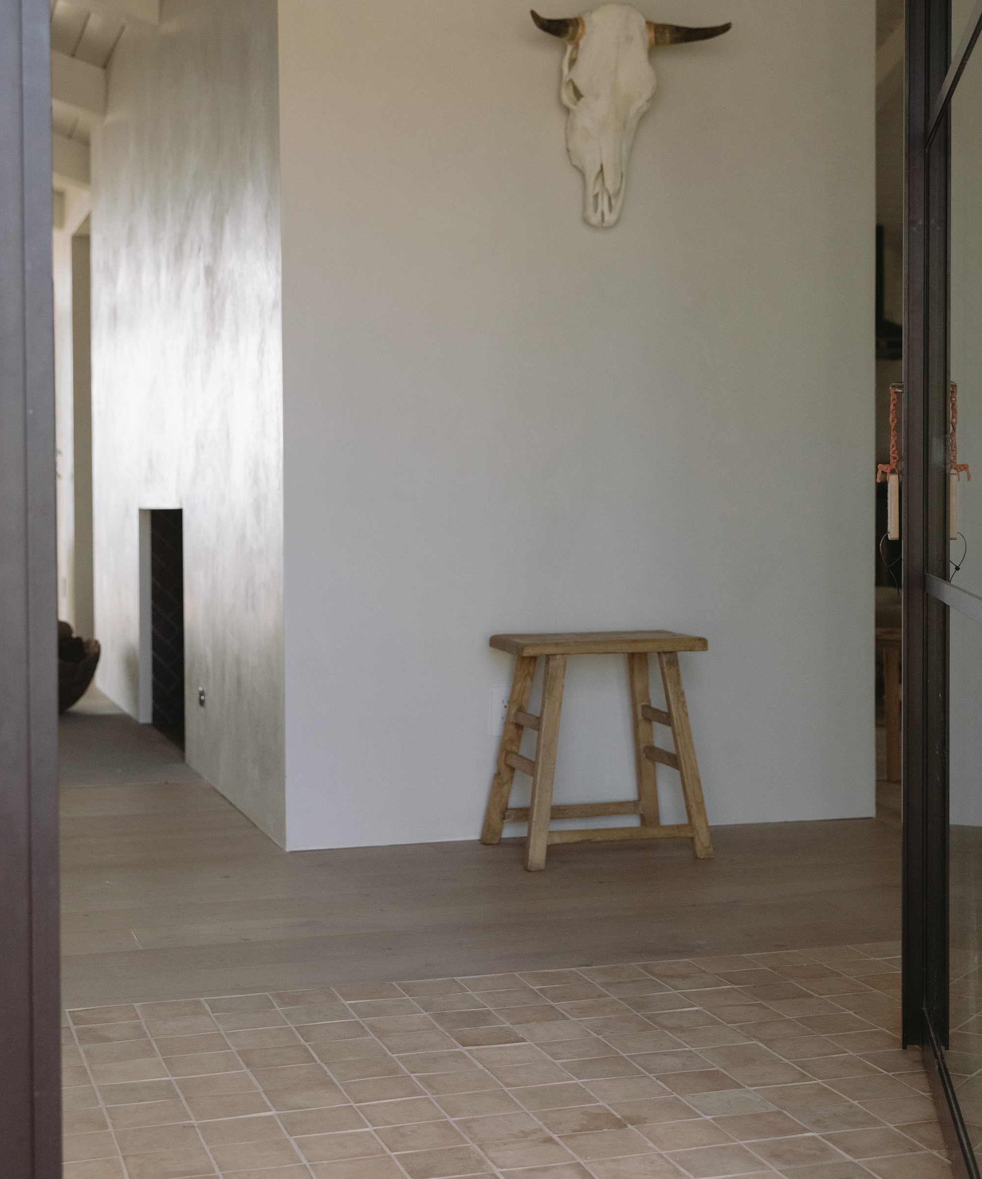 A simple interior with light wood floors, a small wooden stool against a white wall, and a mounted cow skull above. Clé Tile accents the tiled entryway in the foreground, while a fireplace is visible to the left.