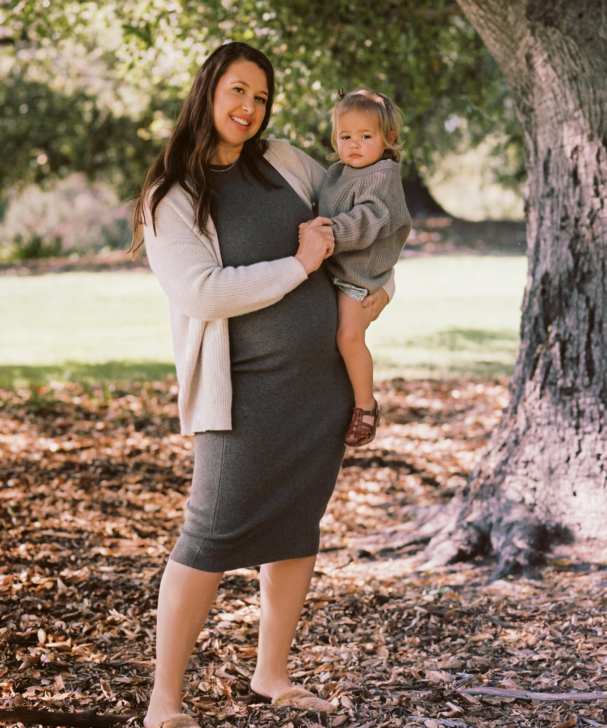 A woman in a gray dress and cream cardigan stands outdoors on leafy ground, holding a young child in a gray sweater. Their classic for life style shines as they look at the camera, surrounded by sunlit trees.
