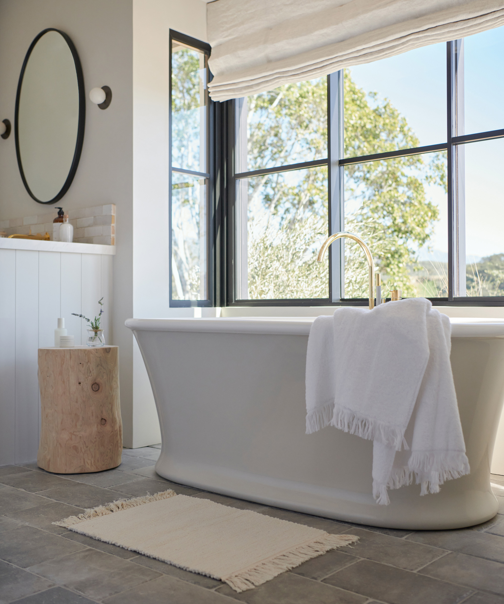Modern bathroom renovation featuring a freestanding bathtub, white towel draped over the side, large windows flooding the space with natural light, a round mirror on the wall, and a wooden stool beside the tub.