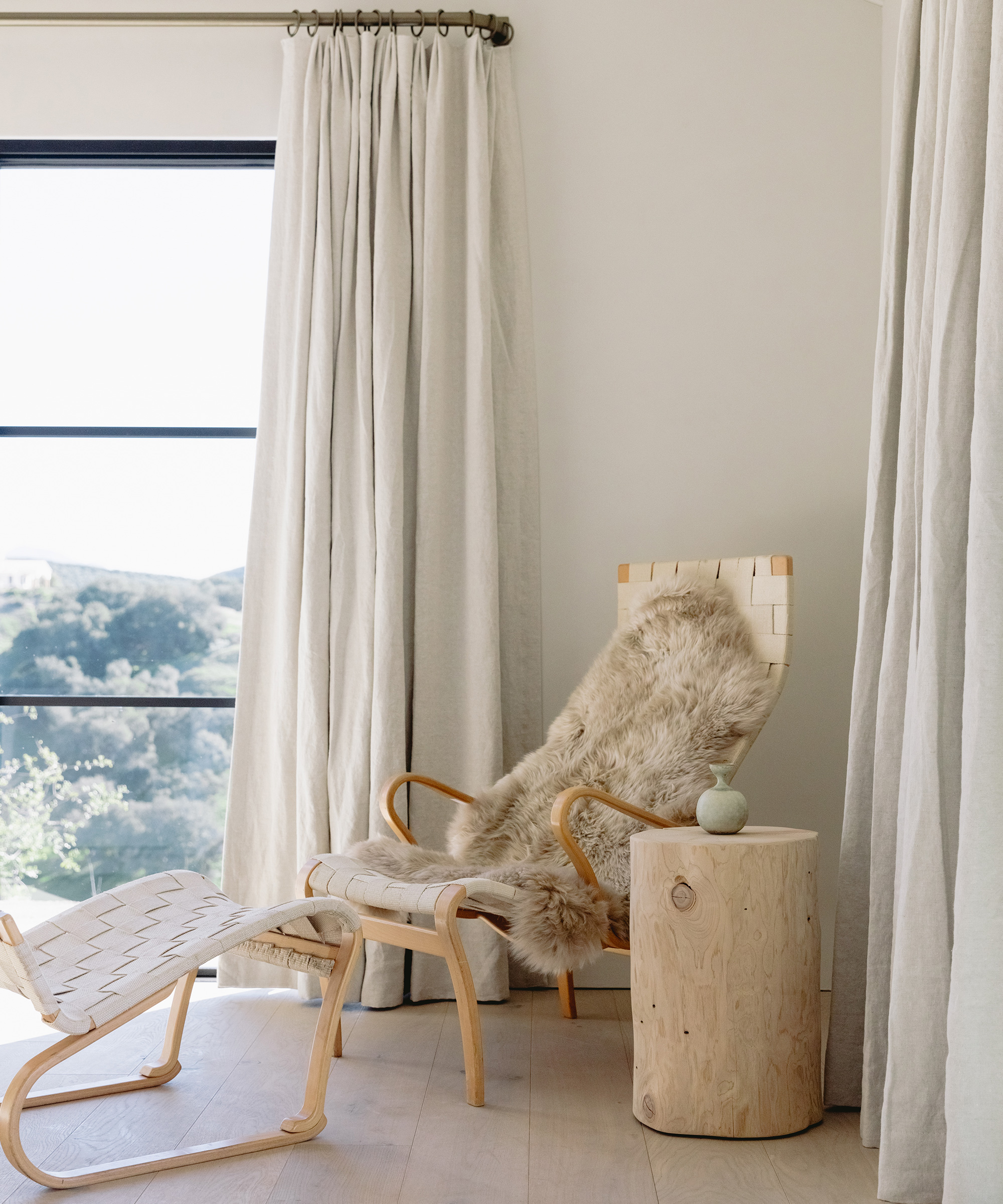 A cozy reading nook, perfect for unwinding after a bathroom renovation, features a modern wooden chair draped with a fur throw, matching ottoman, round side table, and large window with neutral curtains letting in natural light.