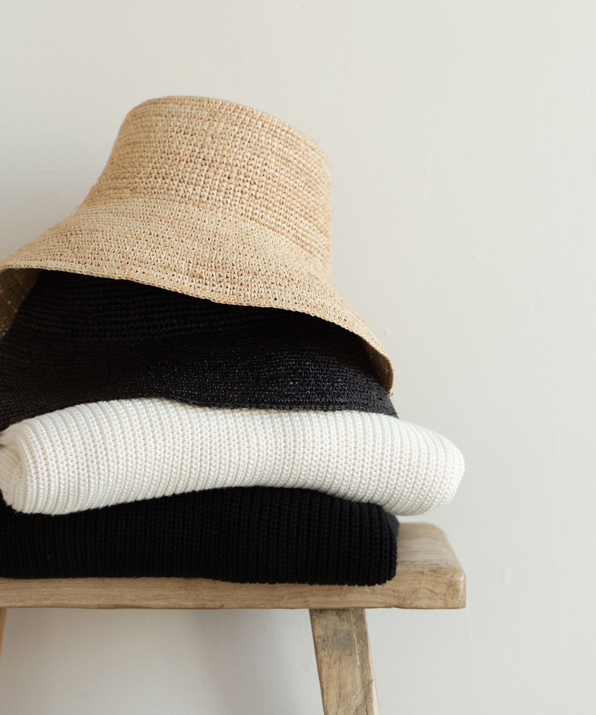 A straw hat rests on top of neatly folded black and white sweaters, placed on a wooden stool against a plain light-colored wall—perfect pieces to dry clean at home for lasting freshness.