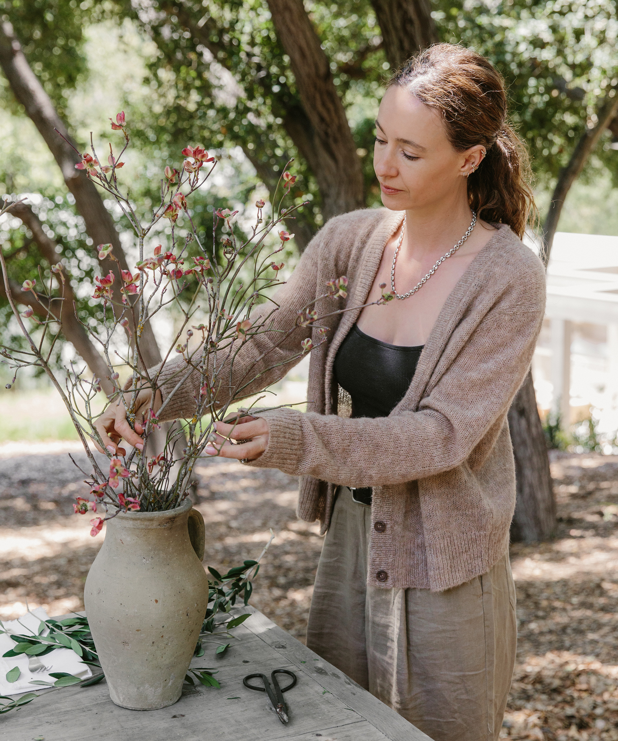 A woman from olive ateliers arranges pink flowers and branches in a beige ceramic vase on a wooden table outdoors, surrounded by greenery and trees. Scissors and foliage rest on the table.