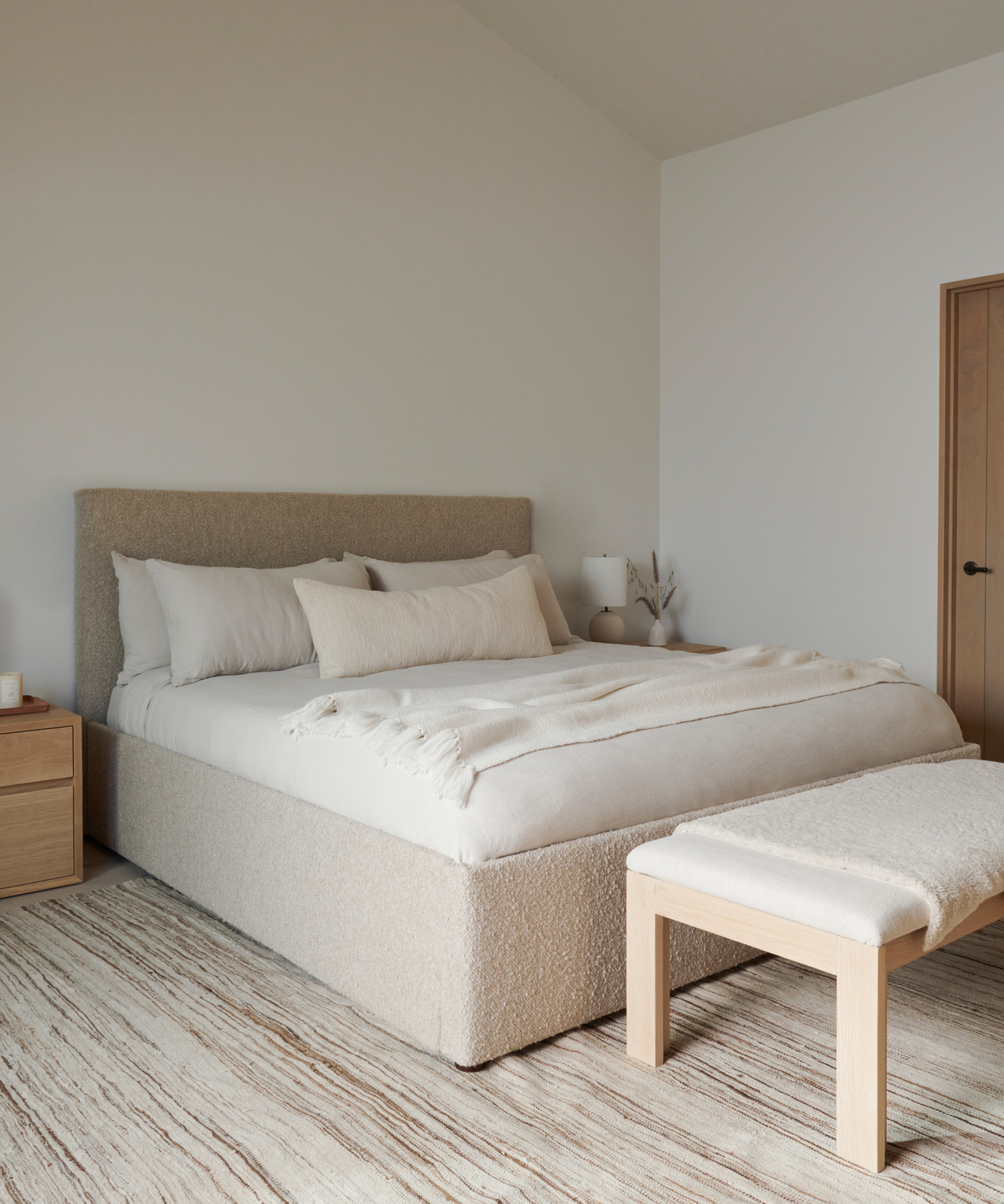 Minimalist bedroom remodel featuring a large upholstered bed, neutral bedding, a wooden bench at the foot, nightstand, and soft natural light—all in beige and white tones for a calm, cozy atmosphere.