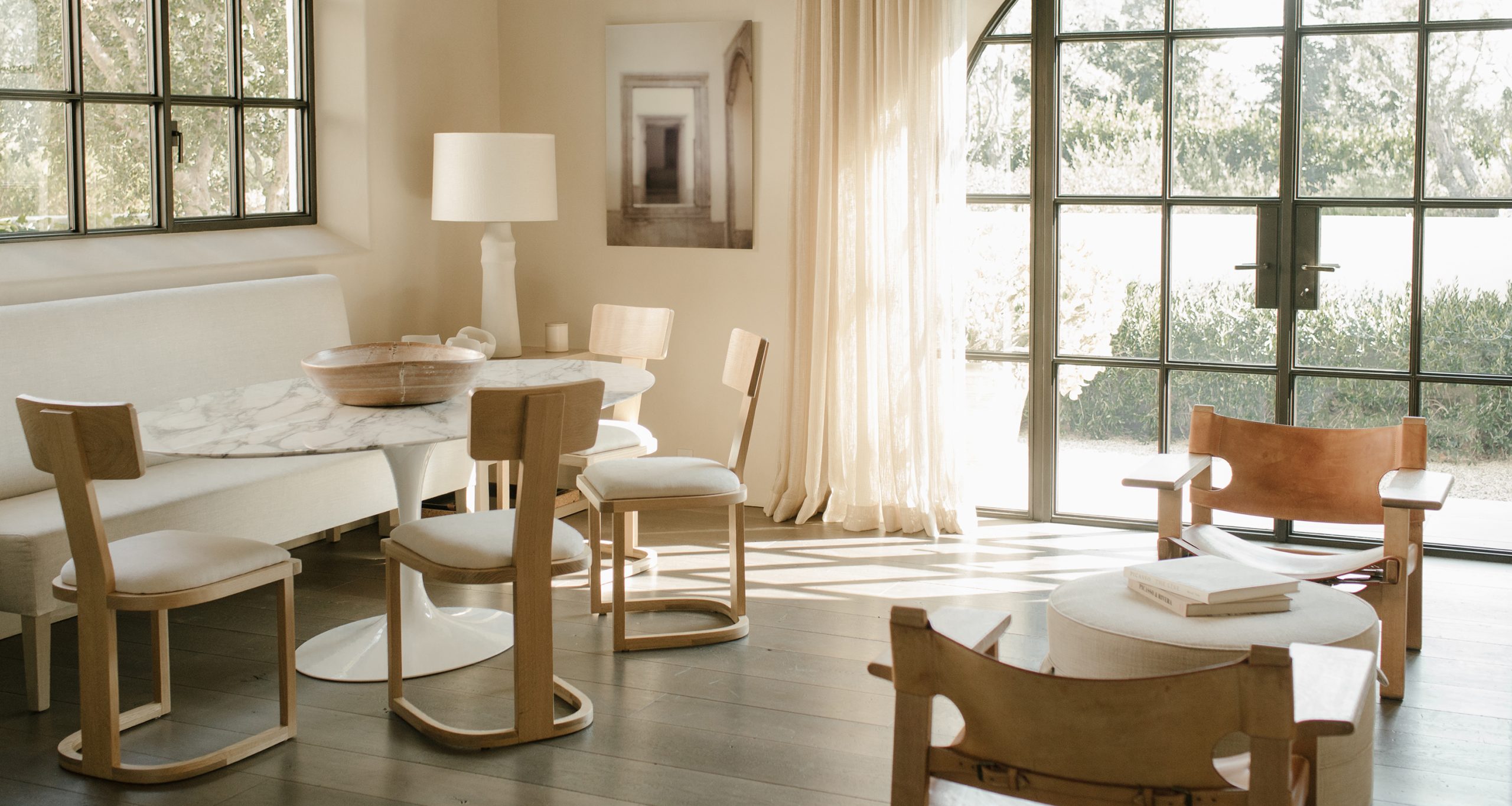 Minimalist living-dining area with wooden chairs, a marble table, white sofa bench, modern lamp, large windows, sheer curtains, sunlight streaming in, neutral color palette, and sleek floors. A framed artwork hangs on the wall.