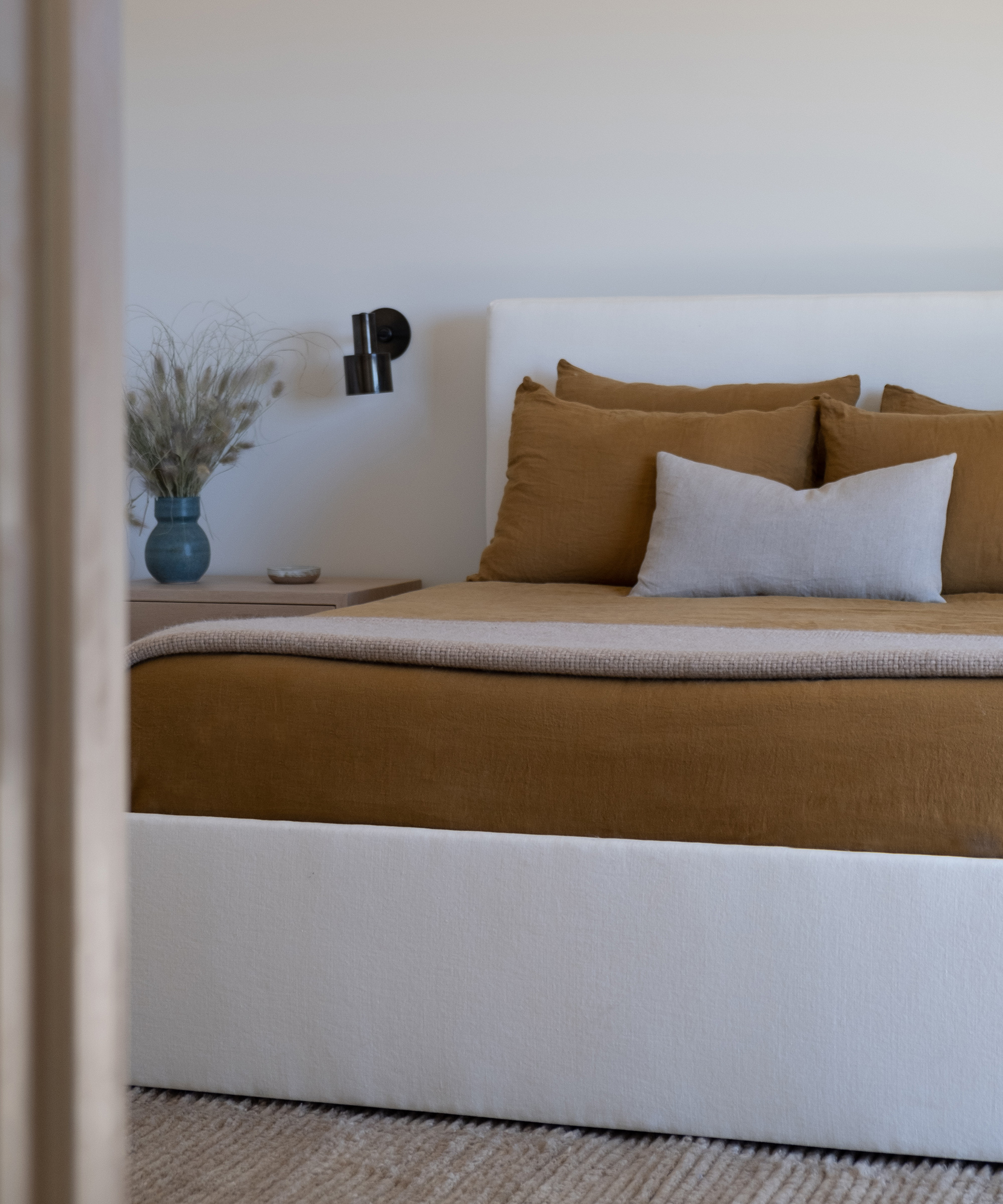 A modern bedroom remodel featuring a white bed frame, tan bedding, and a beige throw blanket. A gray accent pillow and a wooden nightstand with a vase of dried flowers sit beside the bed, with a black wall sconce above.