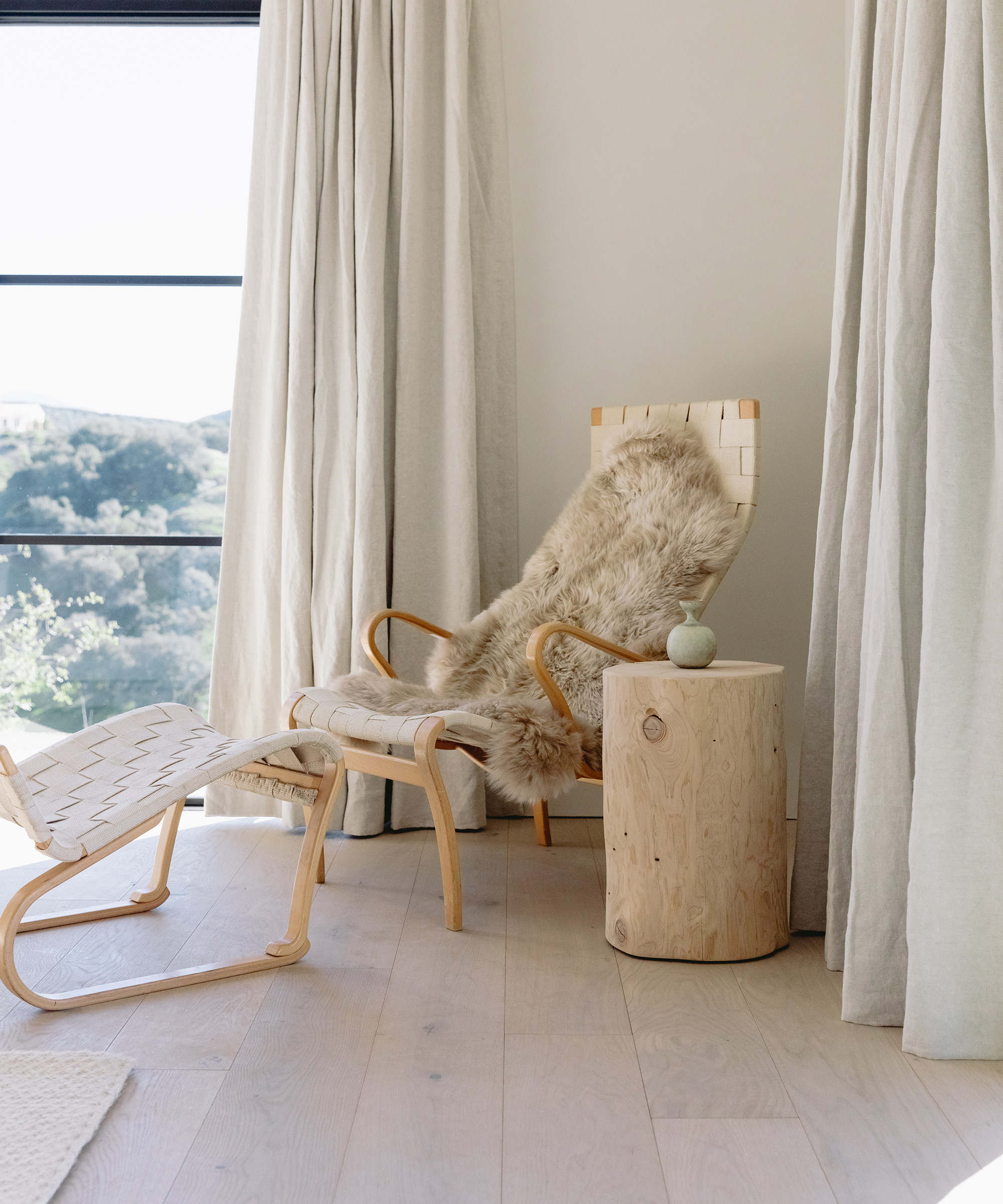 A cozy reading nook with a woven wooden chair, sheepskin throw, matching ottoman, and a round wooden side table holding a small ceramic vase, set by large windows with cream curtains, natural light, and warm wood floors.