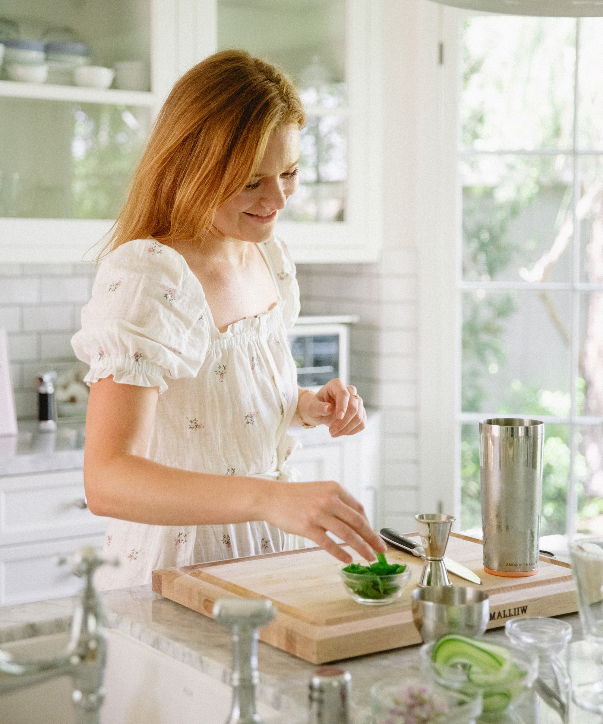 A woman in a white dress stands in a bright kitchen, smiling as she prepares fresh herbs on a wooden cutting board. A cocktail shaker and measuring tools hint at the summer cocktail shes about to craft, while sunlight streams through the window behind her.