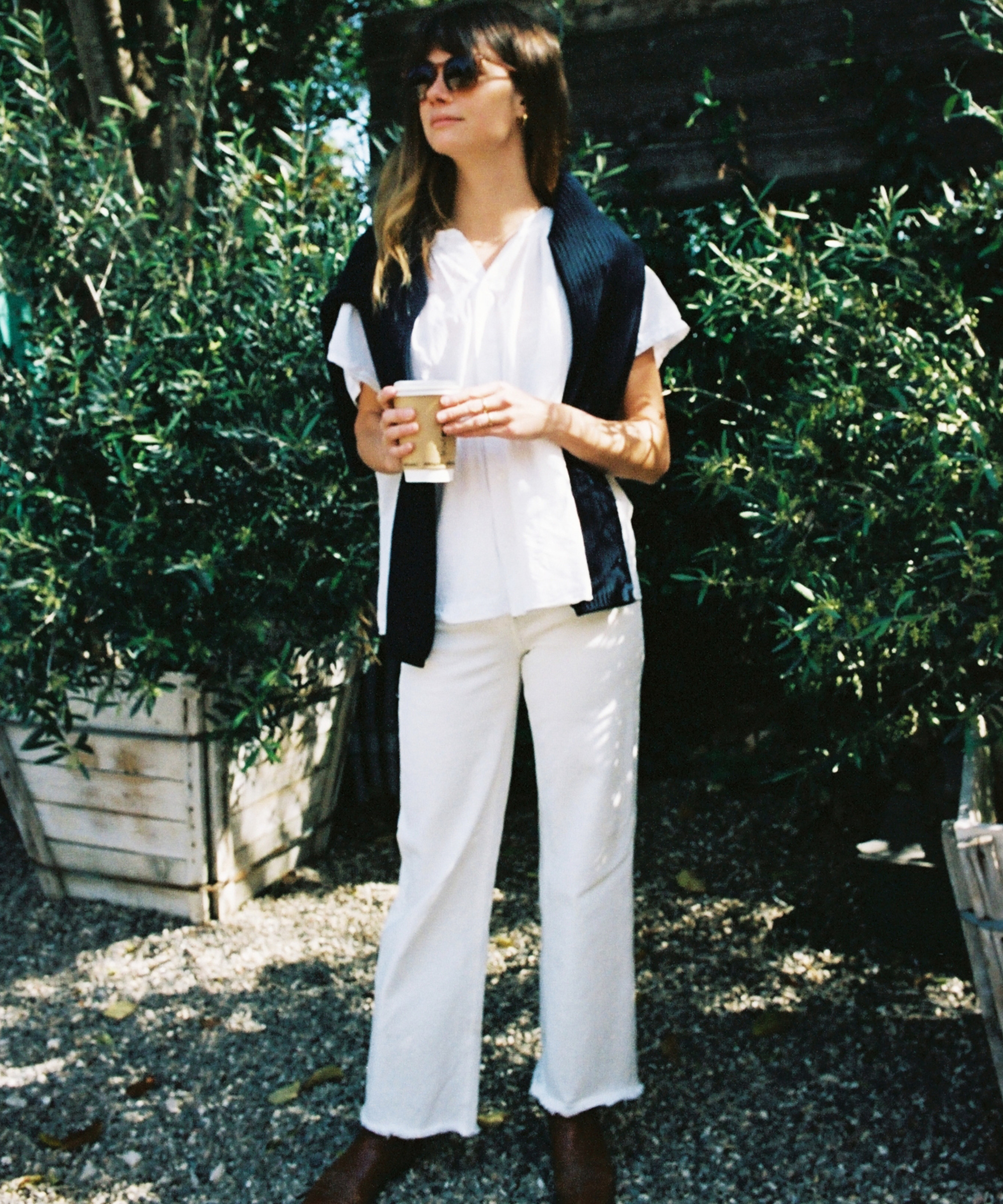 A woman in vintage style sunglasses, a white blouse and pants, brown shoes, and a dark sweater draped over her shoulders stands outdoors with a coffee cup, surrounded by green plants in wooden planters.