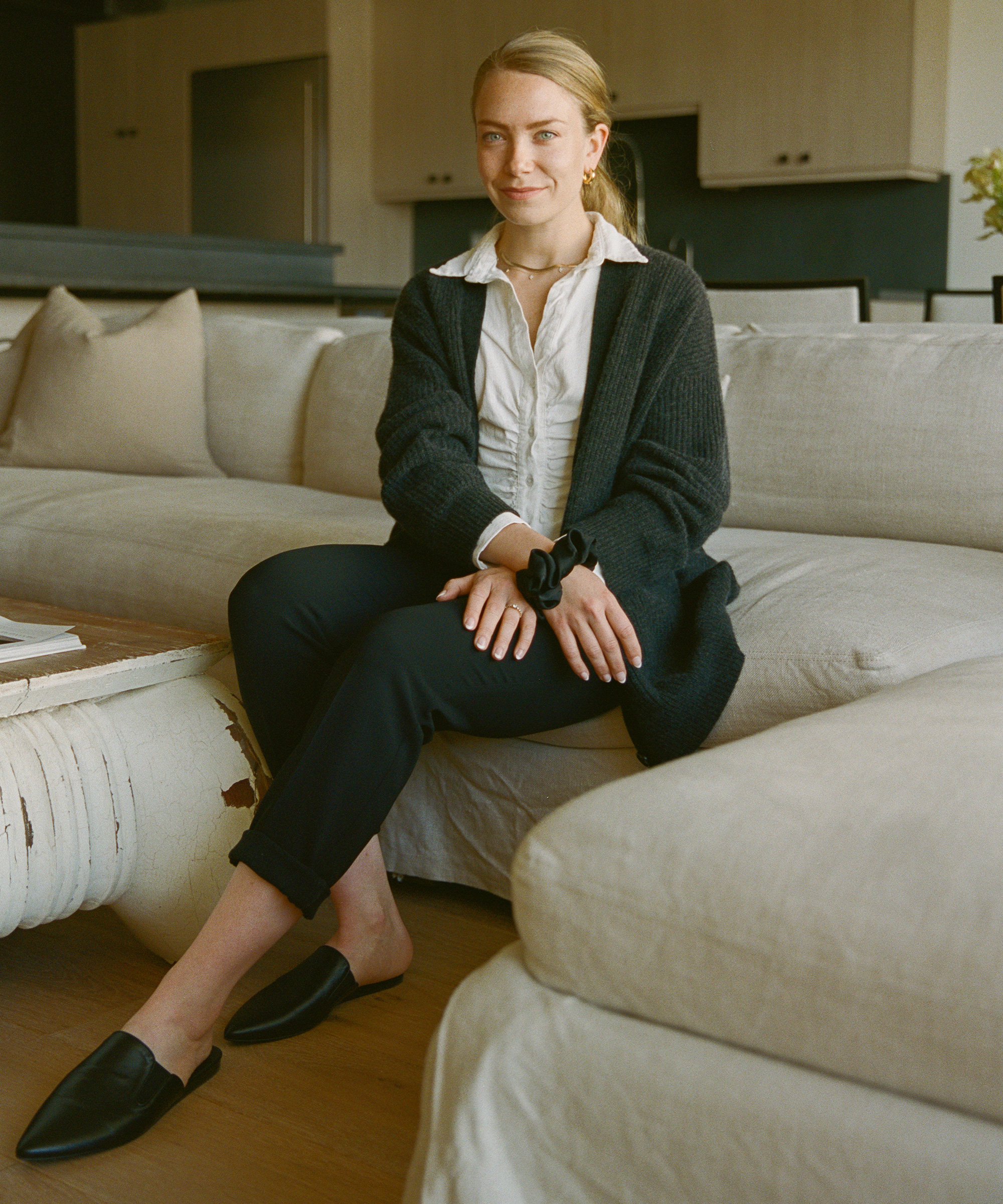 Elisa Baran, with blonde hair tied back, wearing a white shirt, black cardigan, black pants, and black slip-on shoes, sits on a beige couch in a modern living room, smiling slightly at the camera.