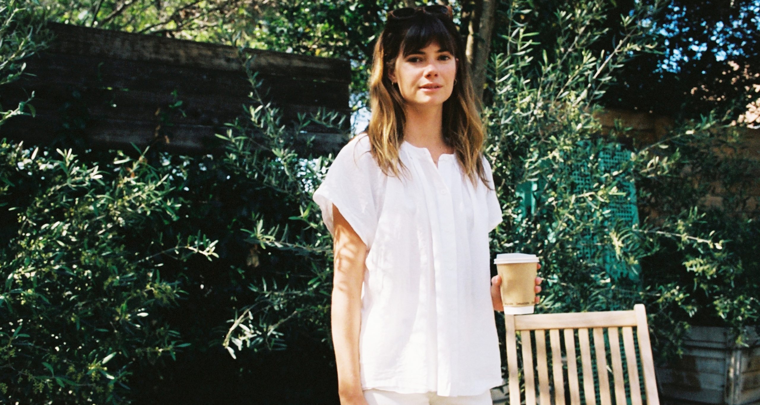 A woman with long brown hair and bangs, dressed in a loose white top, stands outdoors holding a coffee cup. Embracing vintage style, she poses by a wooden chair amid greenery and dappled sunlight.