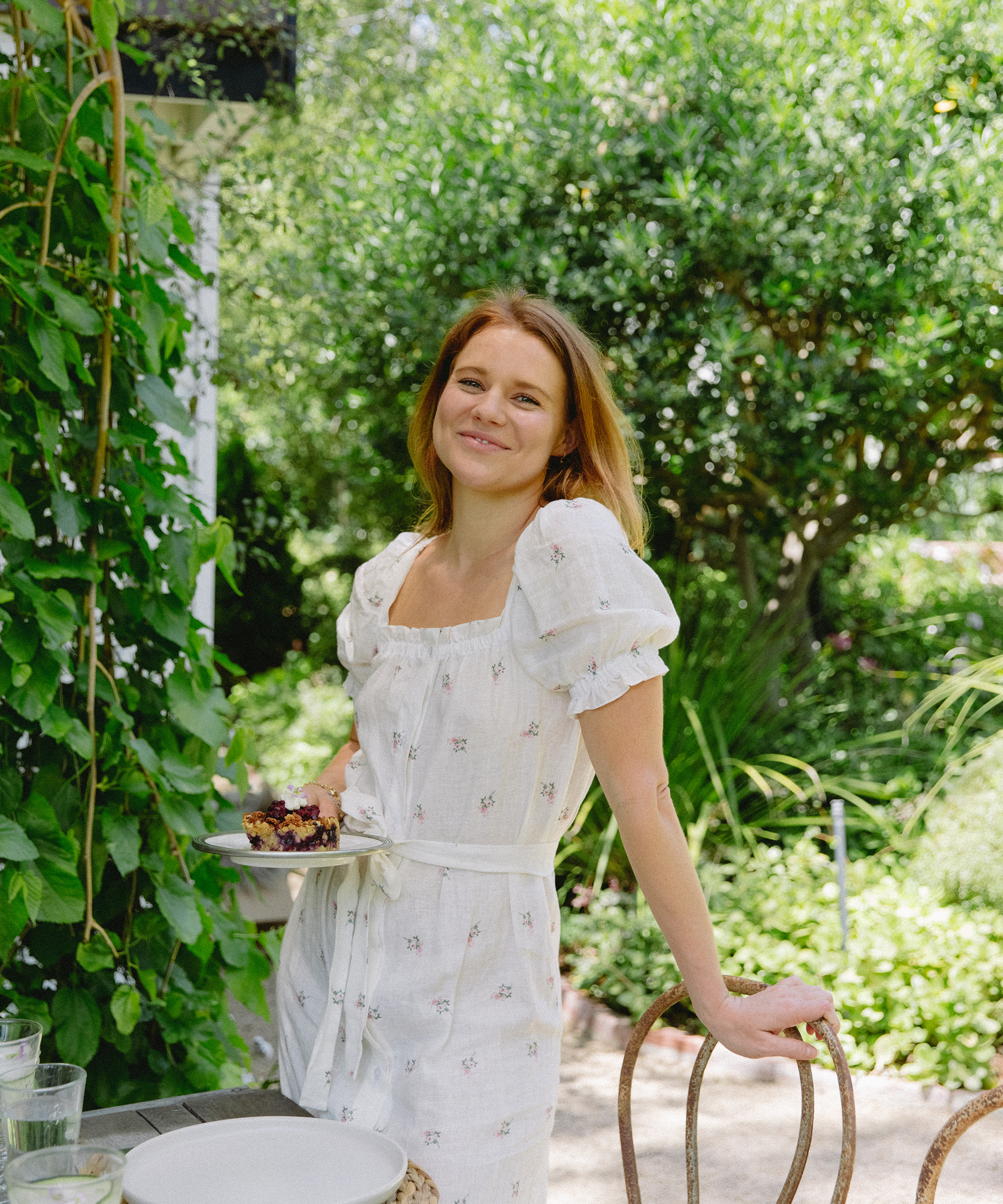 A woman in a white dress stands outdoors by a garden table, smiling and holding a plate of dessert and a refreshing summer cocktail. Lush greenery and sunlight surround her, creating a bright, cheerful atmosphere.