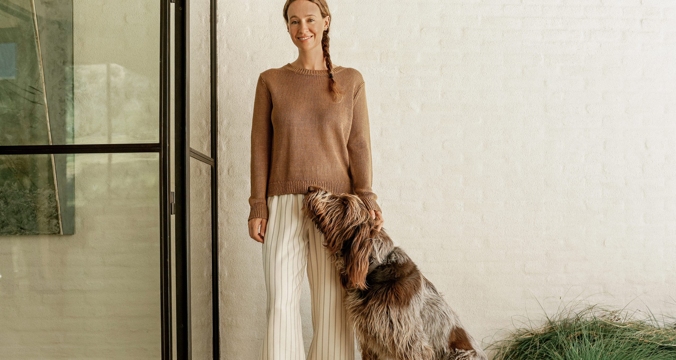 A woman in a brown sweater and white pants, staples of a summer capsule wardrobe, stands indoors smiling as a large, shaggy dog stands on its hind legs, resting its front paws and head against her.