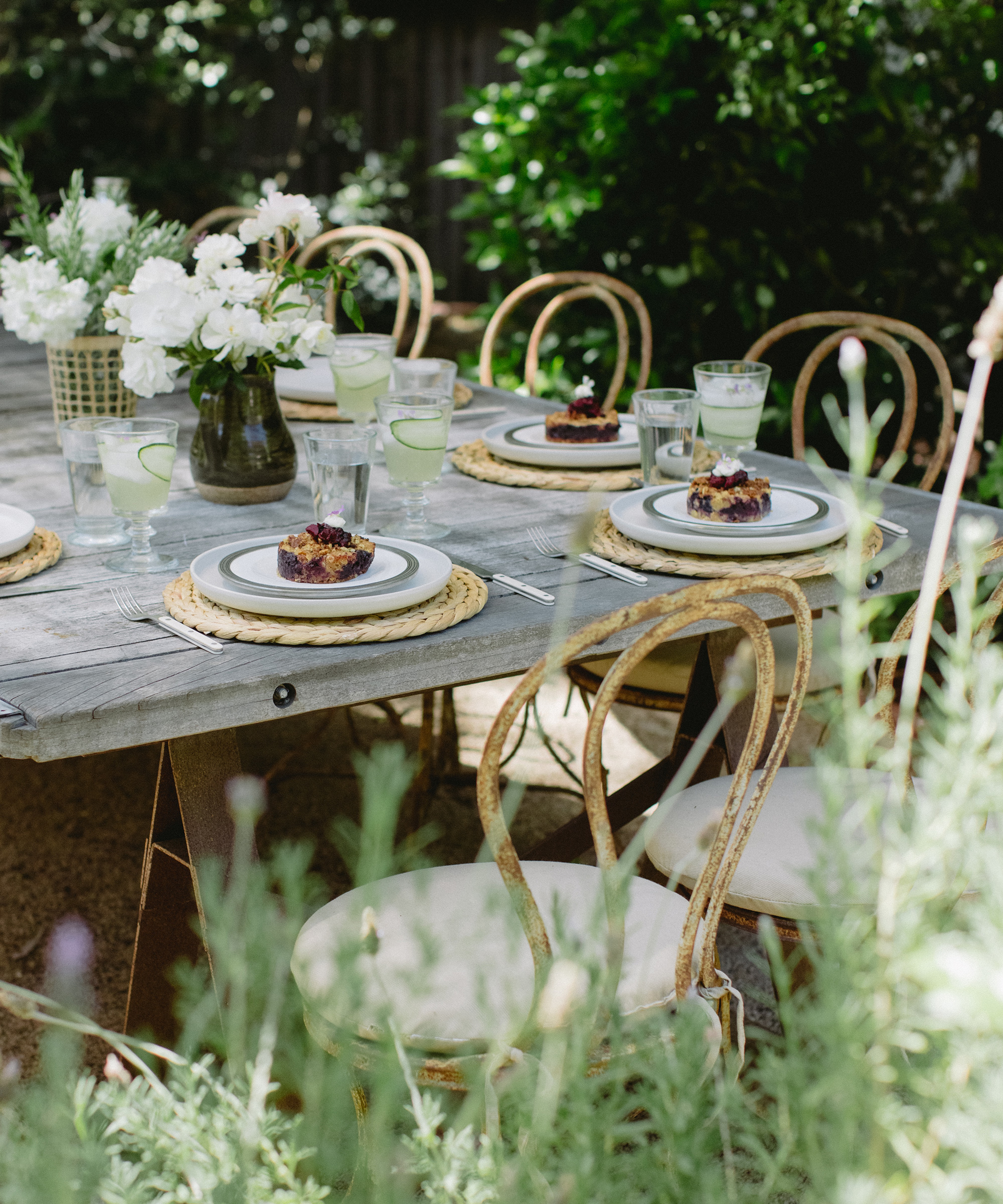 A rustic outdoor table set for four with woven chairs, white plates, floral arrangements, a refreshing summer cocktail, glasses of water with lime, and small berry desserts, surrounded by lush greenery and plants.
