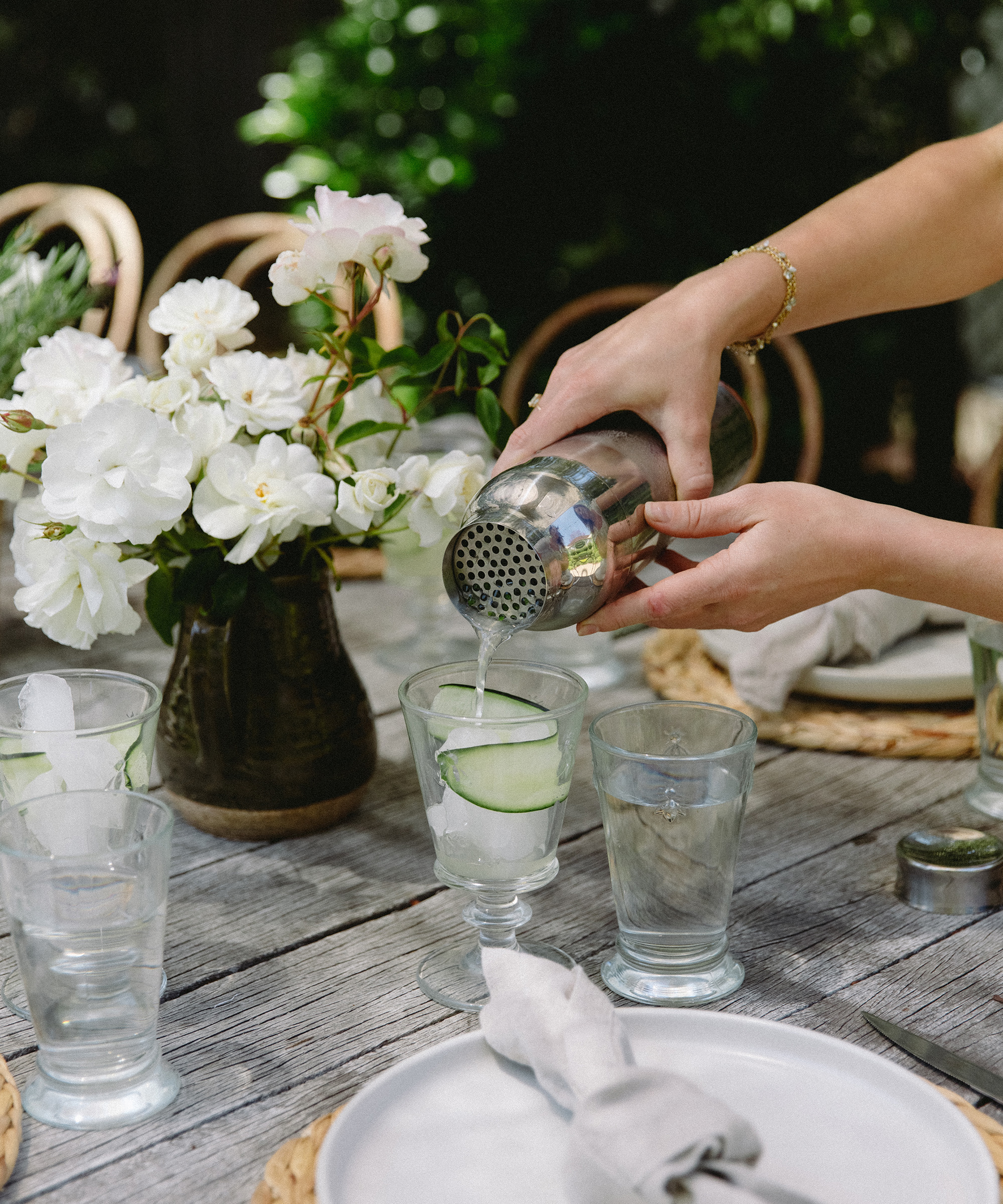 A person pours a refreshing summer cocktail from a shaker into a glass with ice and cucumber on a rustic outdoor table set with plates, napkins, water glasses, and a vase of white flowers.