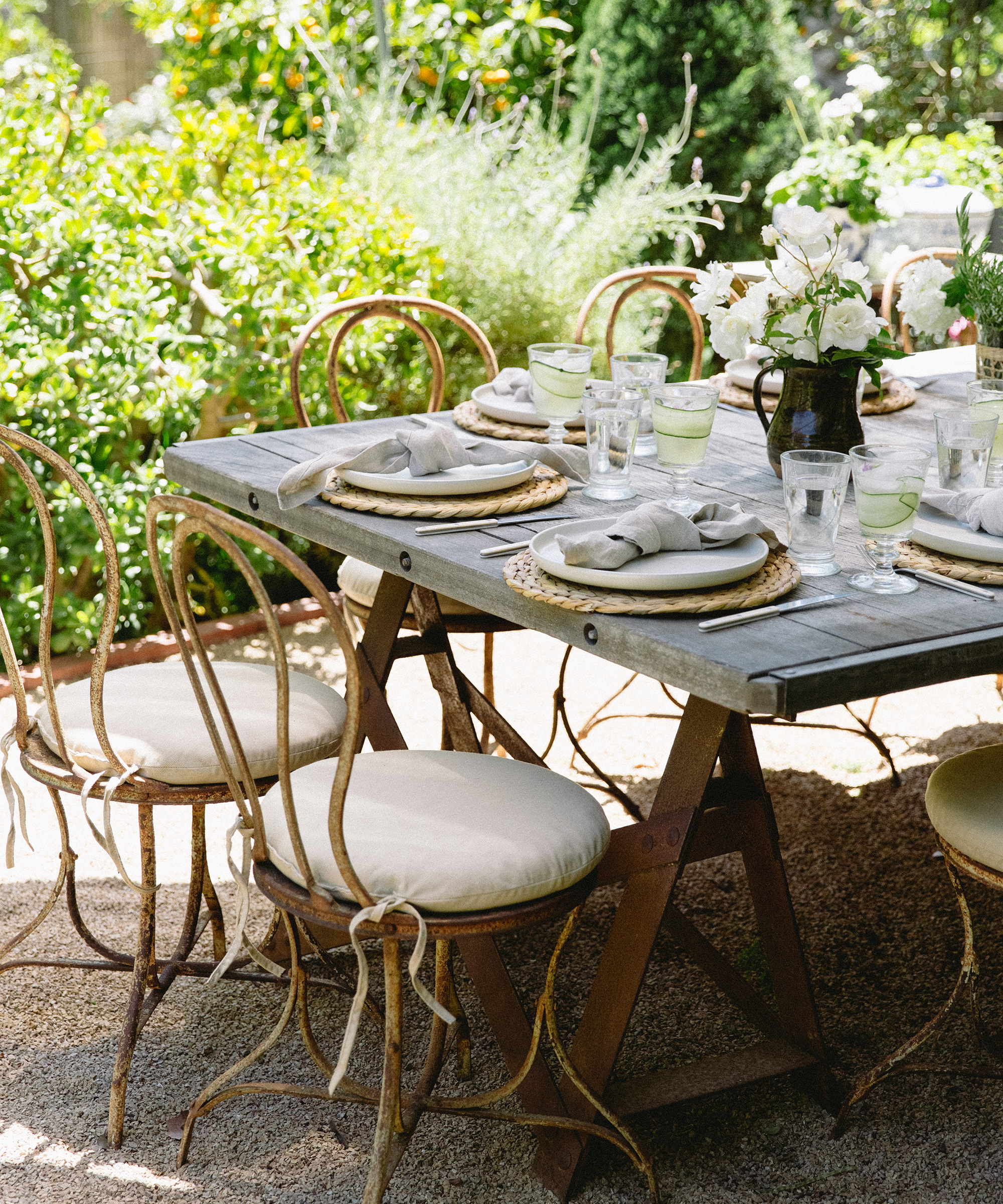 A rustic outdoor dining table set with plates, glasses, and napkins, surrounded by vintage metal chairs with white cushions, in a lush garden setting with greenery, flowers, and refreshing summer cocktails.