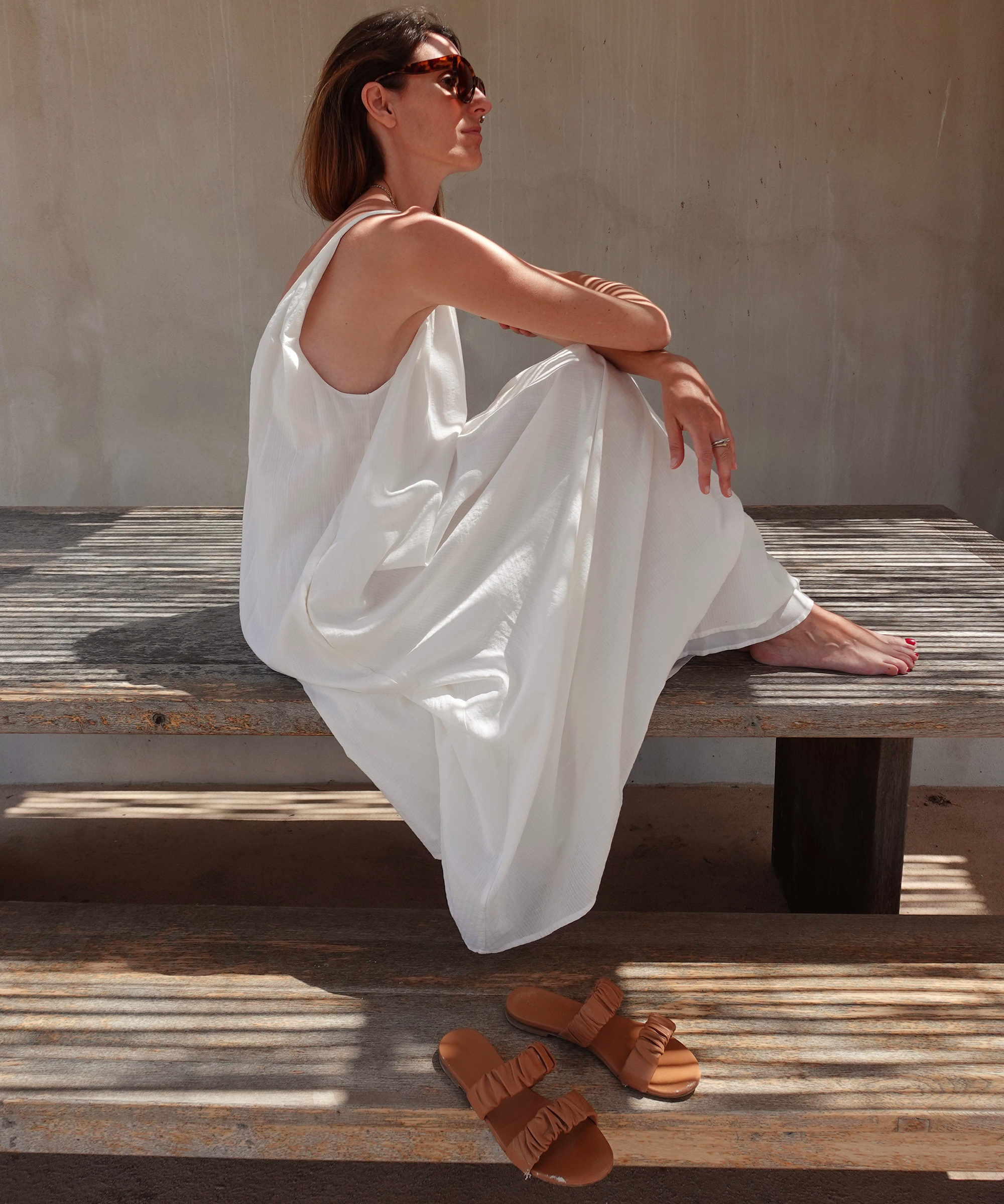 A woman in a flowing white dress sits sideways on a wooden bench, barefoot, with her knees up and arms resting on them. Sunglasses hint at her summer getaway mood, while sunlight creates striped shadows beside sandals on the ground.