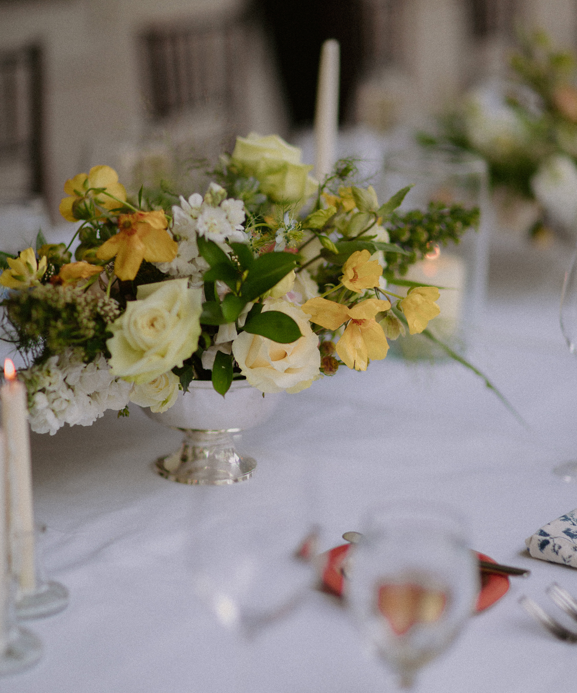 A close-up of a floral centerpiece with yellow and white flowers in a silver vase on a white tablecloth, surrounded by glassware, candles, and blurred chairs—perfect inspiration for wedding tips on elegant table settings.