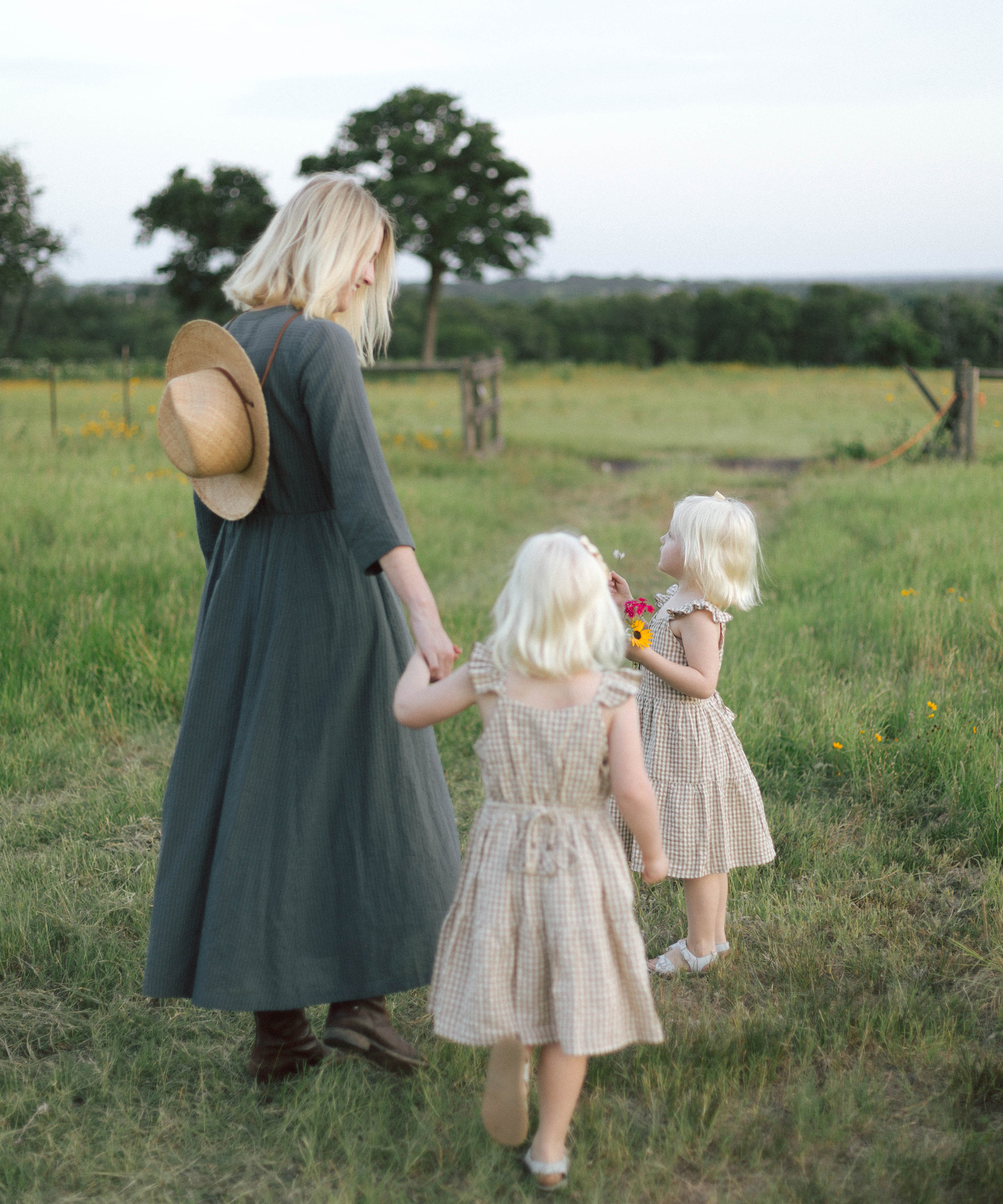 A woman in a long green dress and hat walks hand-in-hand with two young blonde girls in matching dresses through a grassy field, the perfect scene for sharing favorite summer recipes under the trees by a rustic wooden fence.