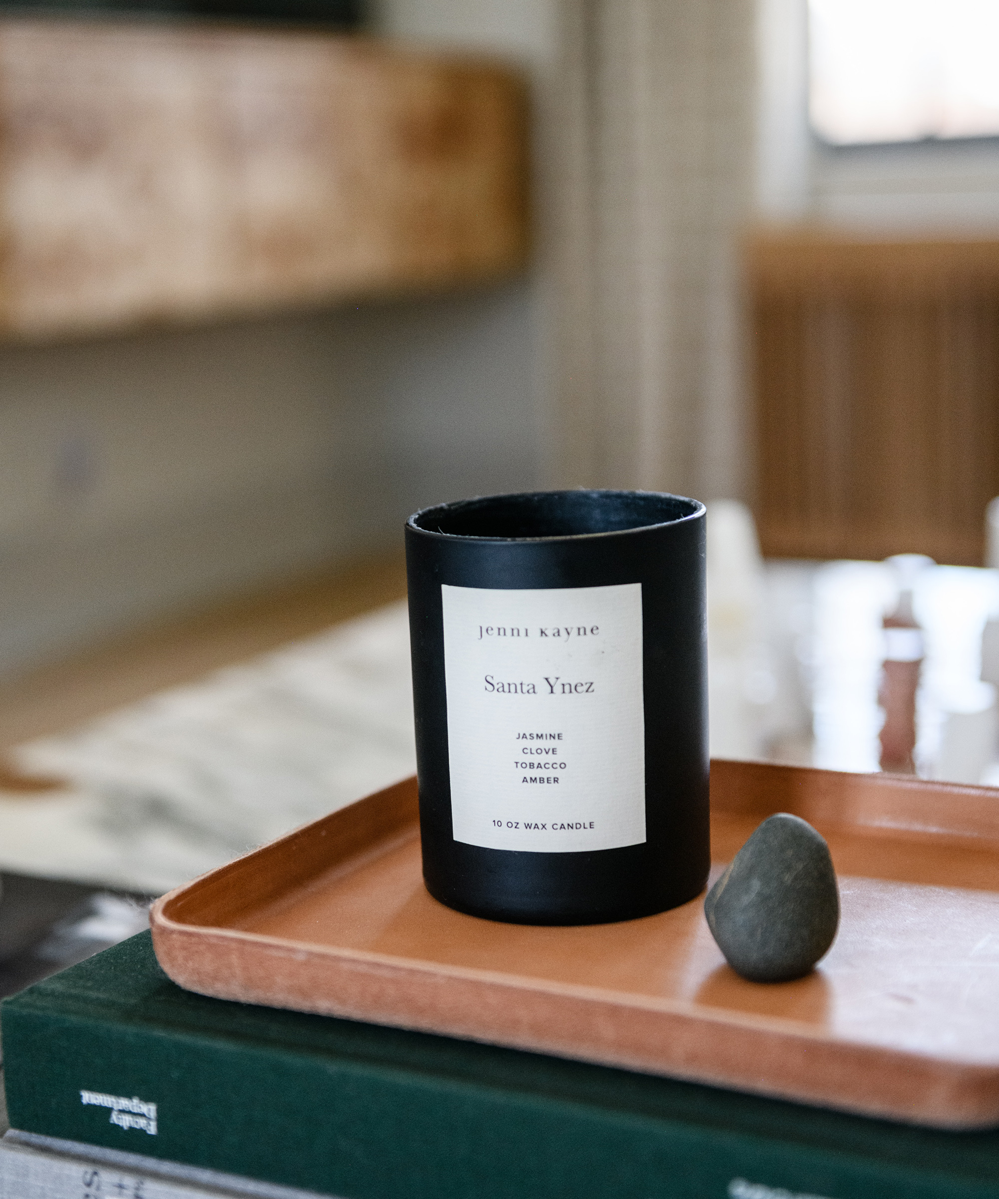 A black candle labeled Santa Ynez by Jenni Kayne sits on a brown tray next to a small gray stone, with Julia Miller’s curated books and blurred furniture in the background.
