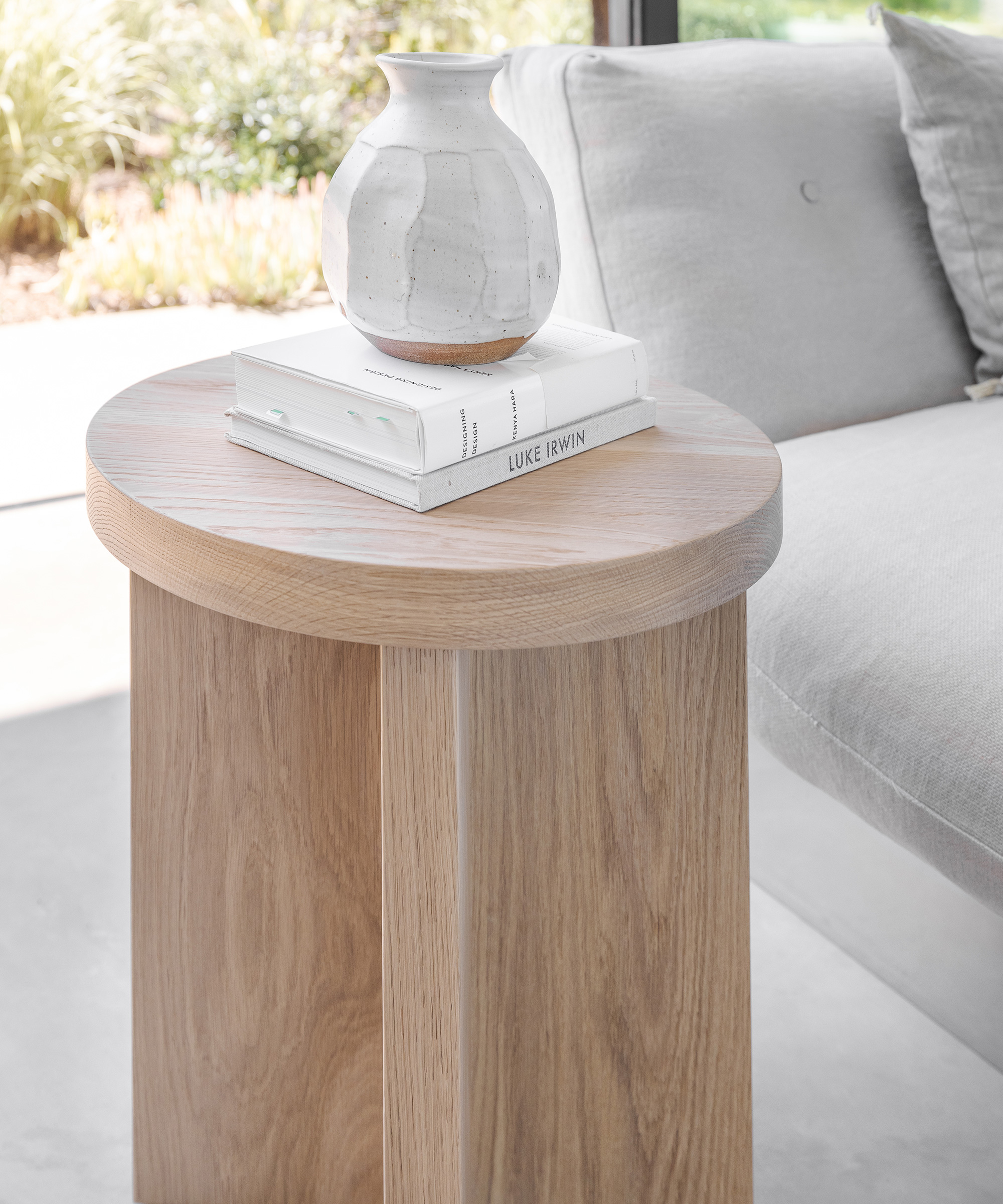 A light wood round side table, inspired by Kristina ONeals modern aesthetic, holds two stacked books and a white ceramic vase. The table sits next to a light gray sofa in a bright living space with sunlight cascading through the window.