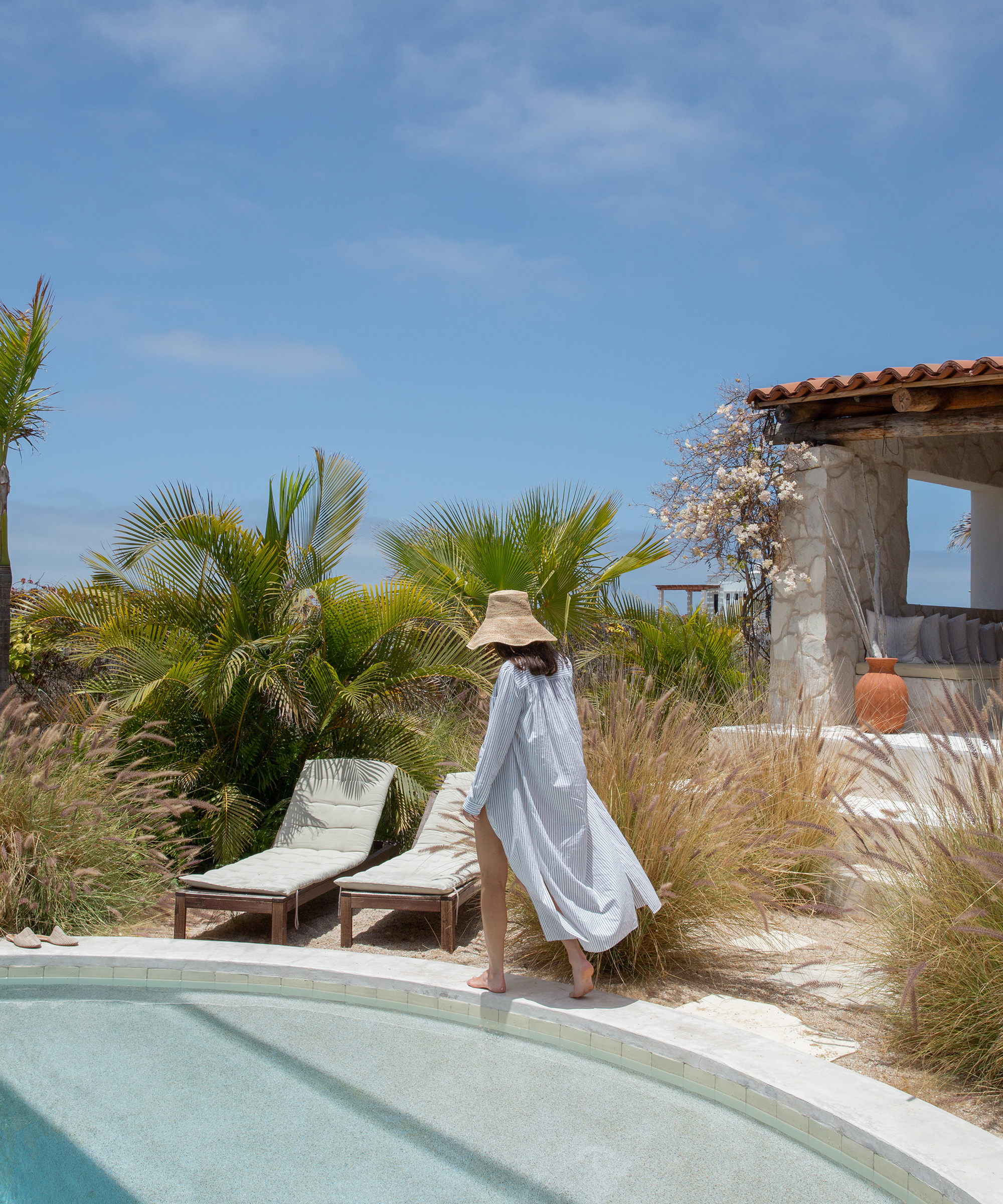 A woman in a sun hat and white cover-up walks beside a pool toward lounge chairs, surrounded by palm trees and grass, with a stone cabana in the background under the blue Todos Santos sky.