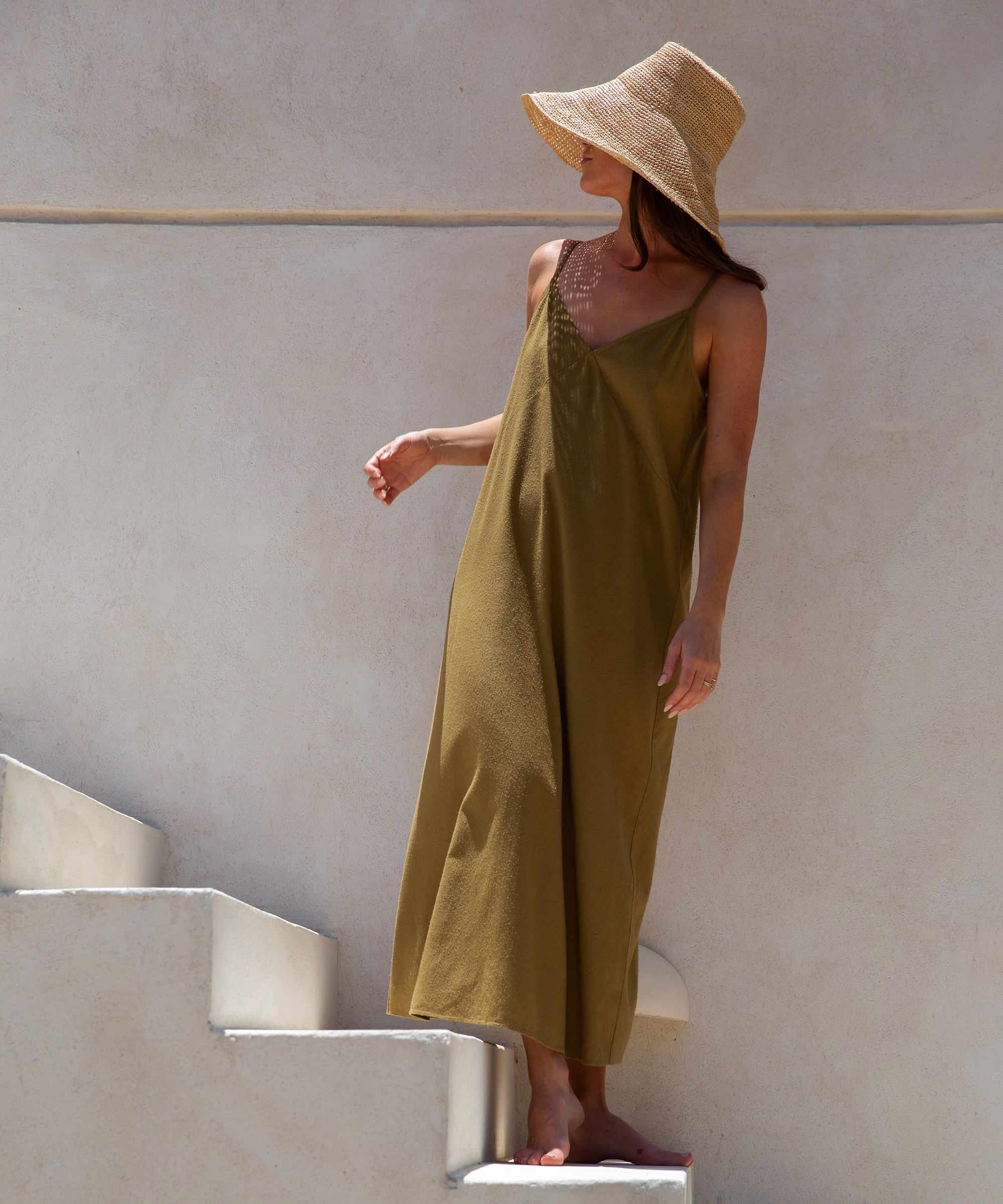 Woman in a wide-brimmed straw hat and loose olive-green dress stands barefoot on white steps against a light beige wall in Todos Santos, looking to her left with one hand by her side and the other slightly raised.