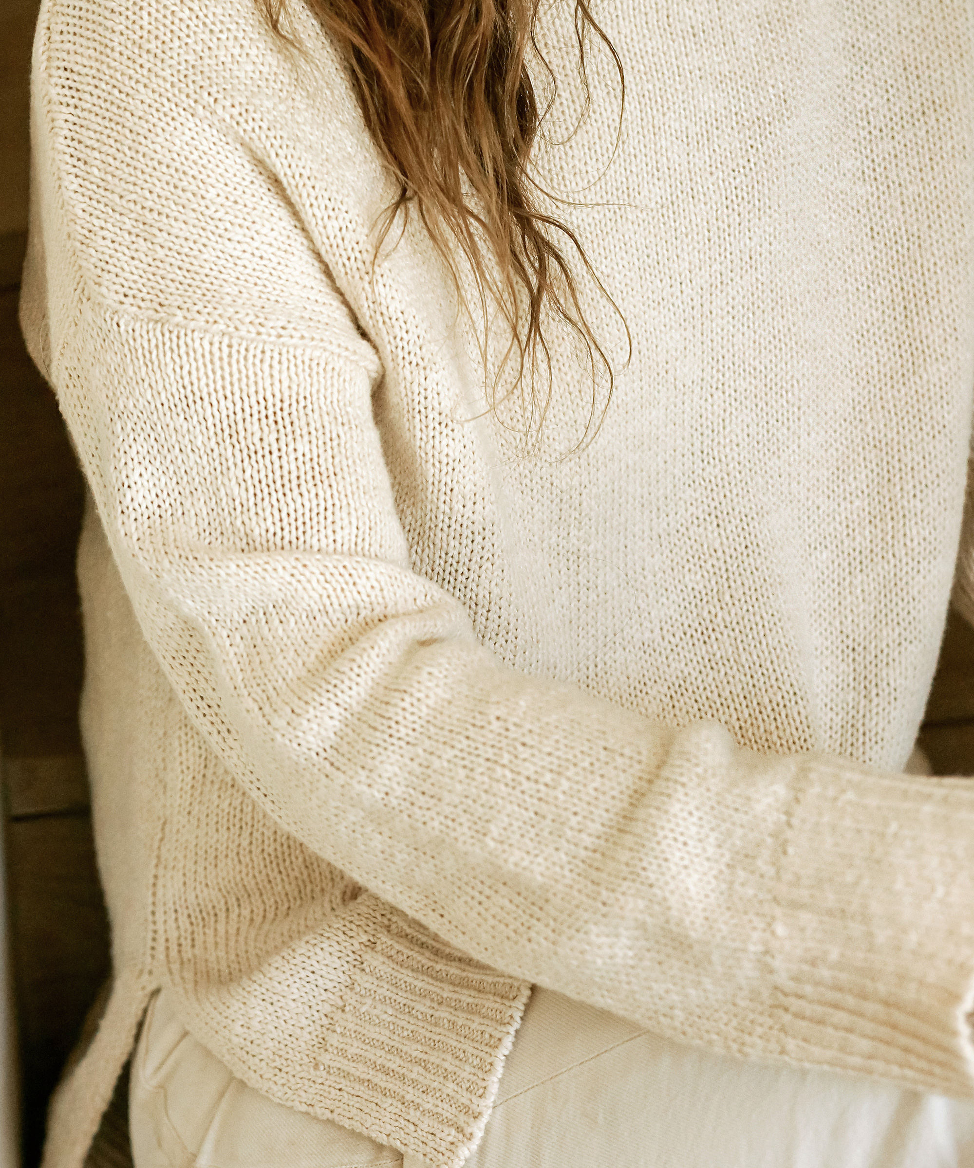 A person wearing a cream-colored knit sweater with long sleeves sits indoors. Their wavy hair partially covers their shoulder, and their face is out of the frame. Despite being a cozy look, it pairs effortlessly with relaxed summer outfits.
