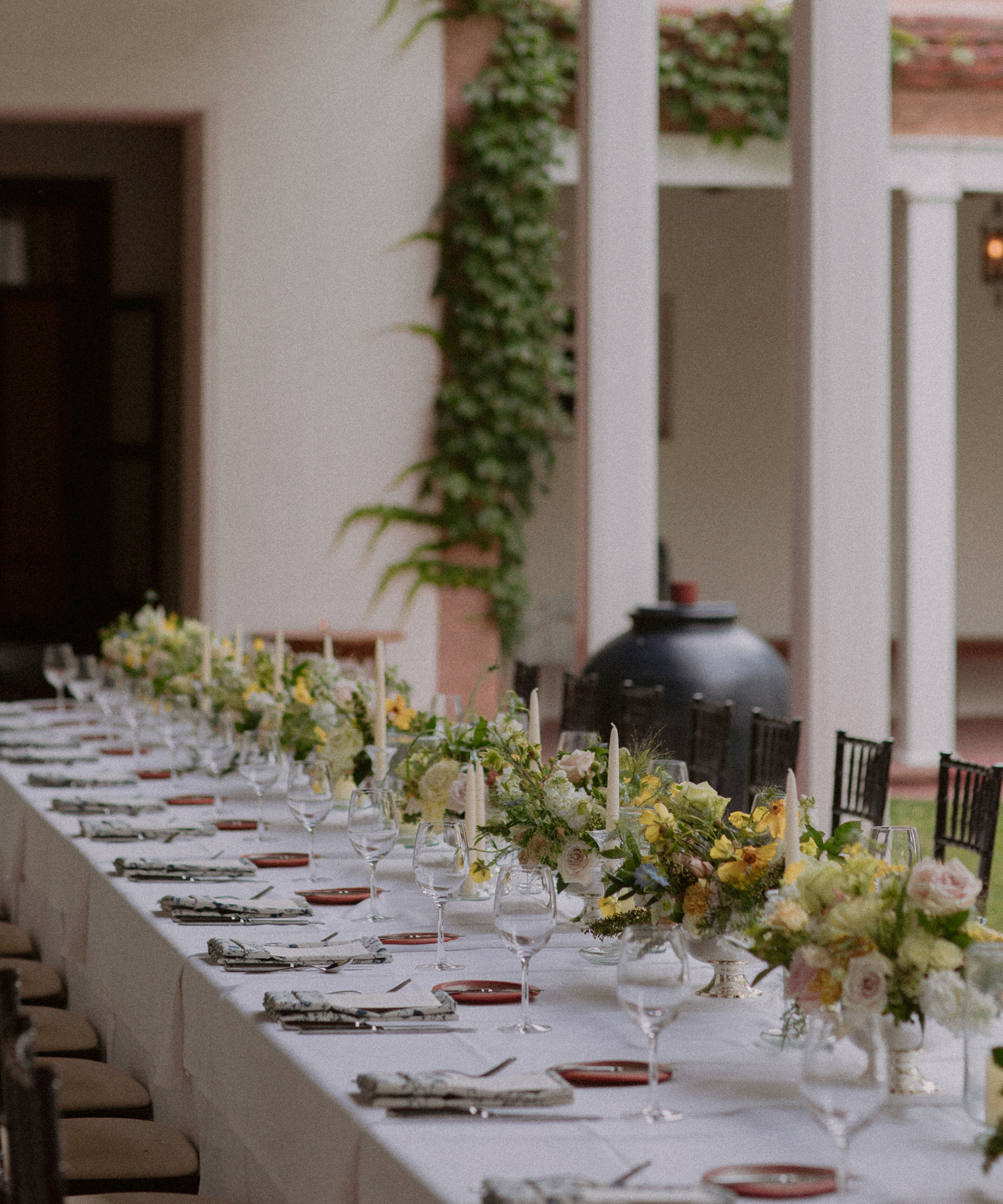 A long, elegantly set dining table with white linens, glassware, plates, and floral centerpieces is arranged outdoors under a covered patio with greenery climbing a column—perfect inspiration for wedding tips on hosting stylish outdoor receptions.