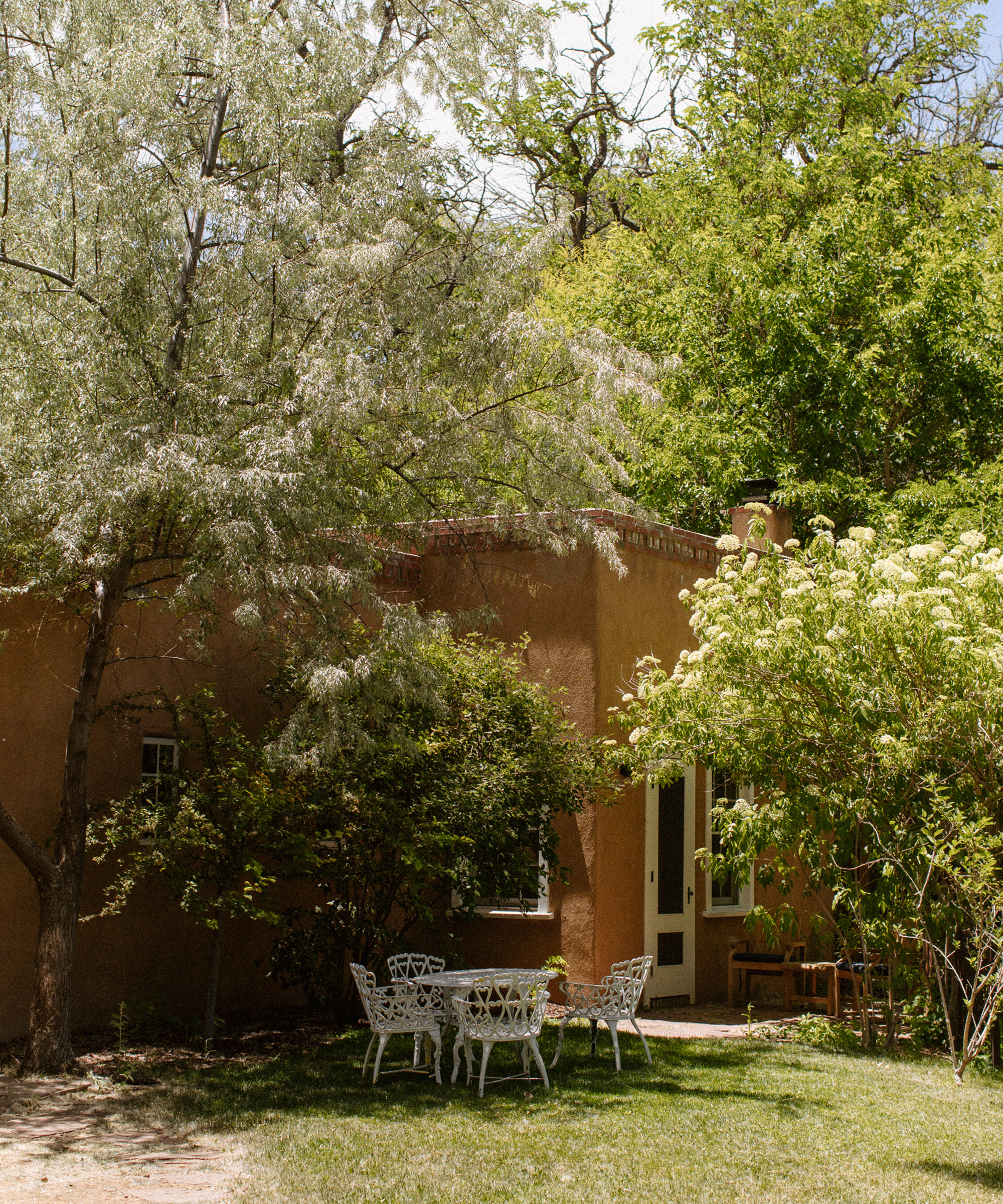 A small, adobe-style house is surrounded by lush green trees and bushes. In the shaded grassy yard, a white metal table and chairs offer a peaceful outdoor setting—perfect for intimate gatherings or sharing wedding tips beneath sun-dappled leaves.
