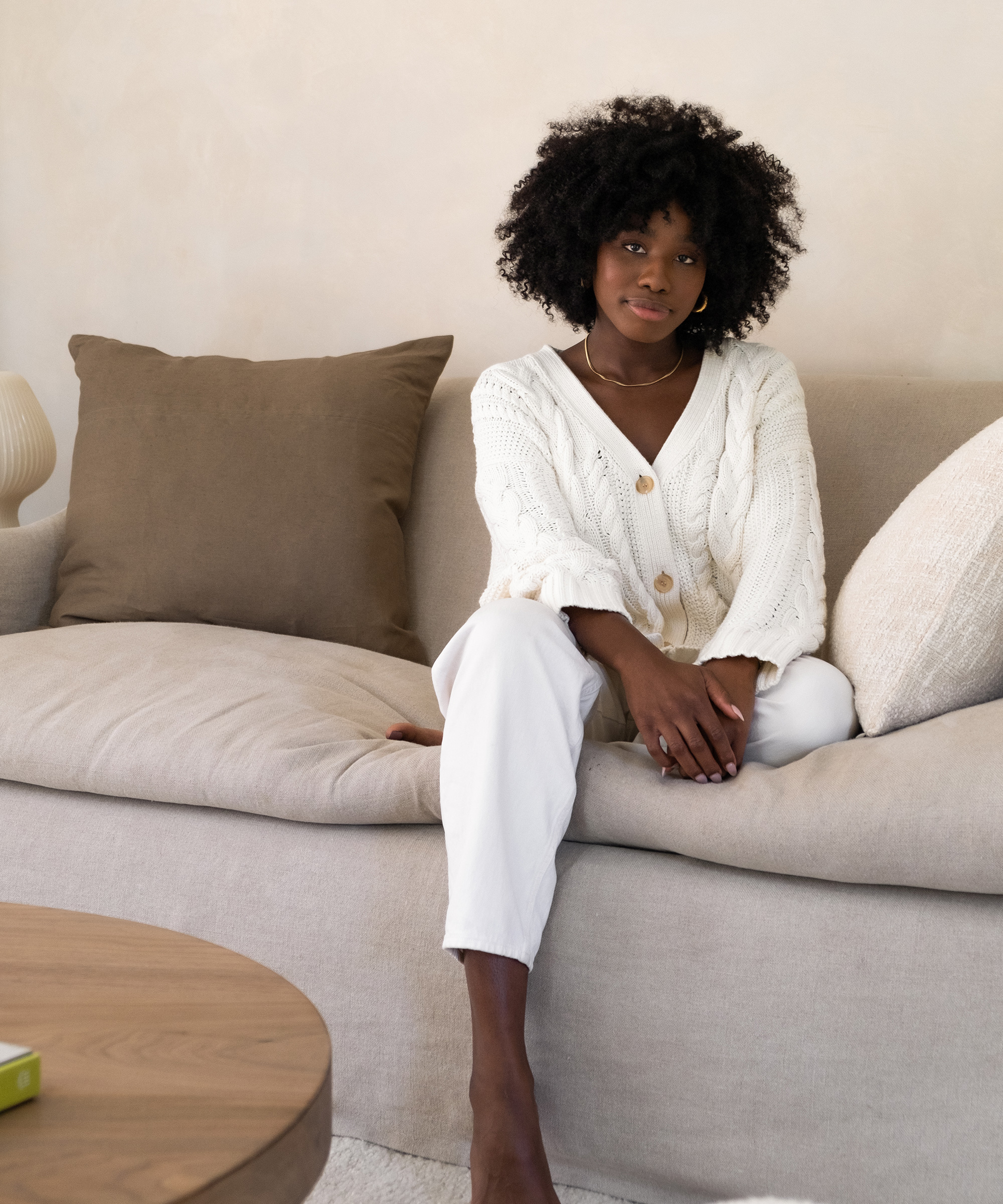 A woman with curly hair sits on a beige sofa with brown and cream pillows, wearing a white knit sweater and white pants. She looks at the camera with a relaxed expression in a cozy, minimal living room after a recent room renovation.