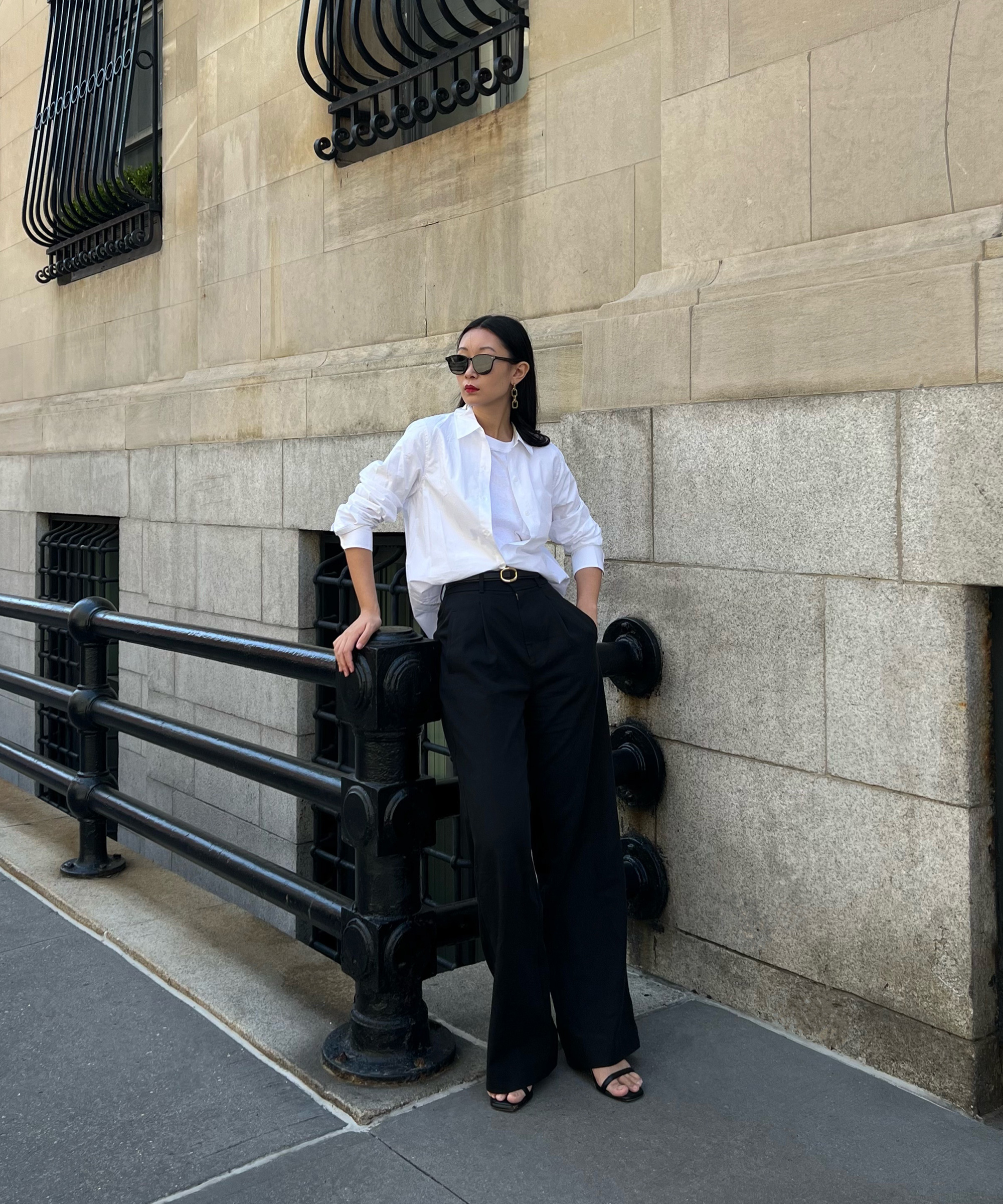 A woman showcasing her transitional wardrobe stands on a city sidewalk, wearing sunglasses, a white button-up shirt, and high-waisted black pants as she leans against a black railing before a beige stone building.