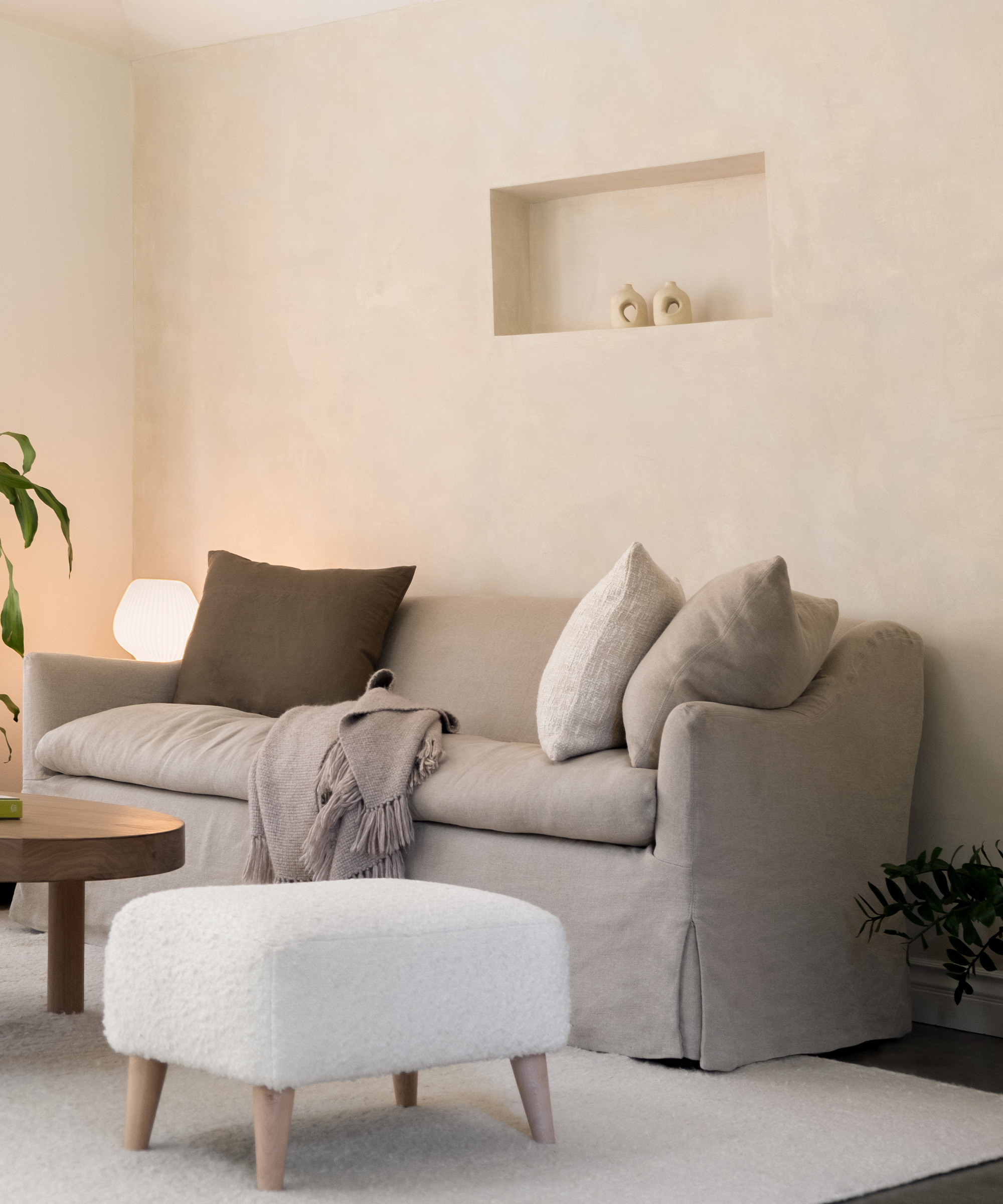 A cozy living room designed after a thoughtful room renovation, featuring a beige sofa, neutral cushions, soft throw blanket, white ottoman, round wooden coffee table, glowing lamp, green plants, and simple decor on a recessed wall shelf.