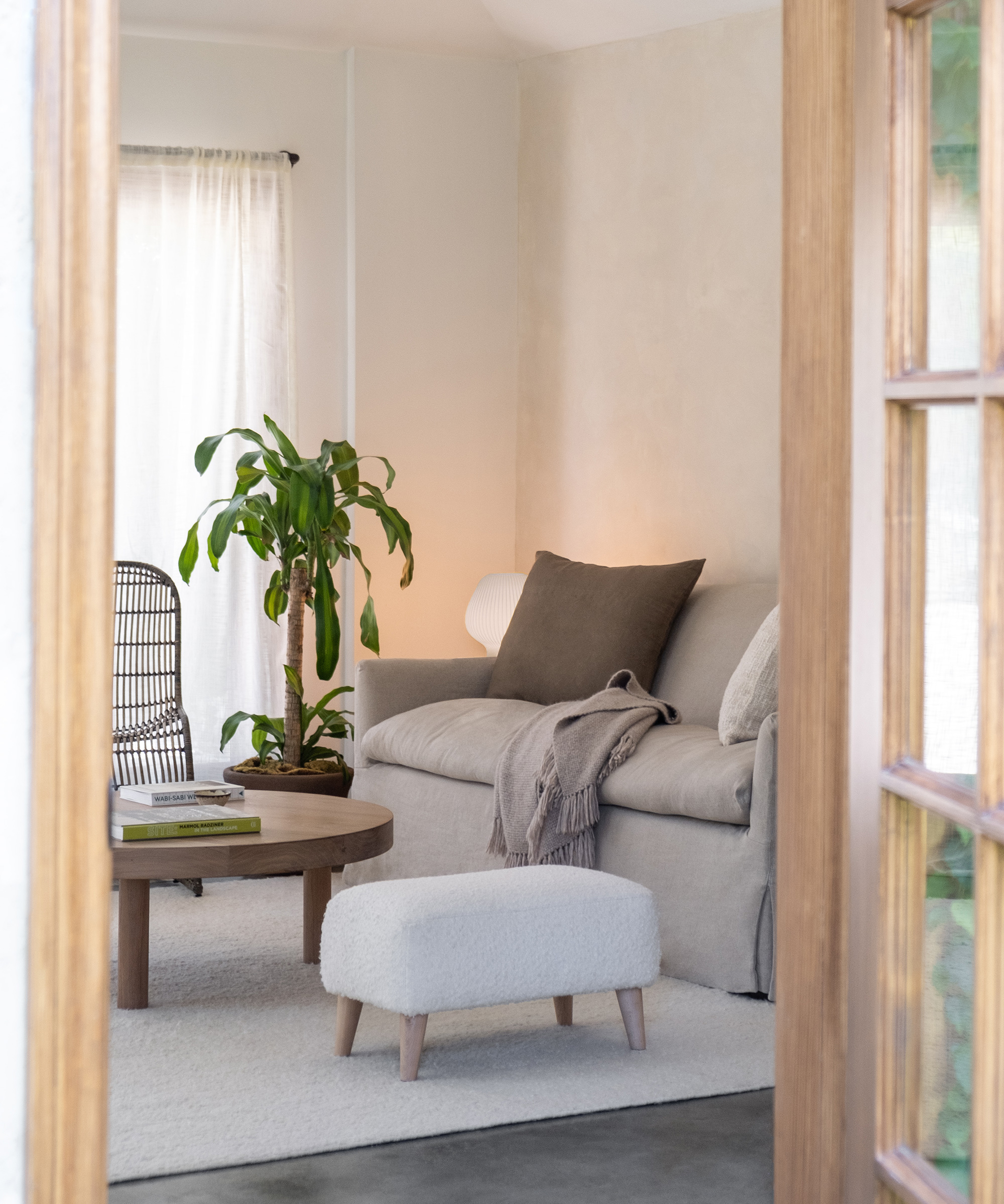 A cozy living room renovation features a beige sofa, throw blanket, small round coffee table, potted plant, upholstered footstool, and natural light streaming through a sheer curtain—all viewed through a wooden door.