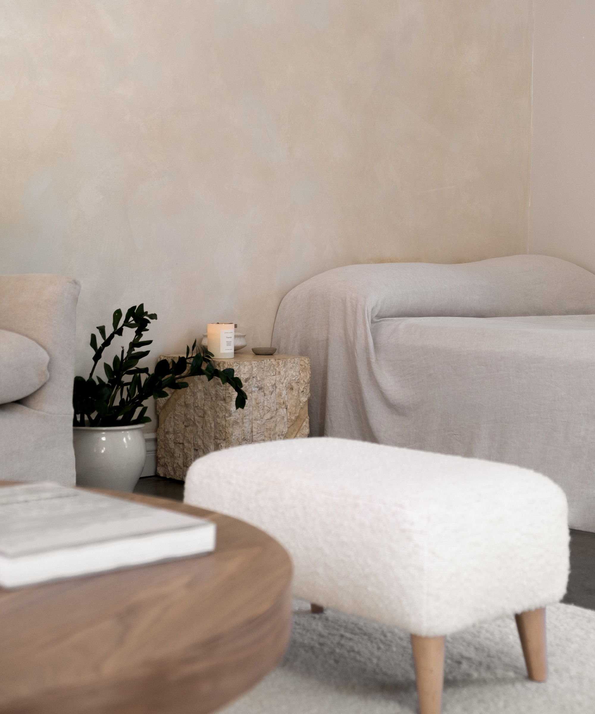 A minimal, cozy living room with light-colored furniture, a wooden coffee table, and a textured ottoman—perfect inspiration for your next room renovation. A stone side table holds a candle and book, while a potted plant rests against a neutral wall.