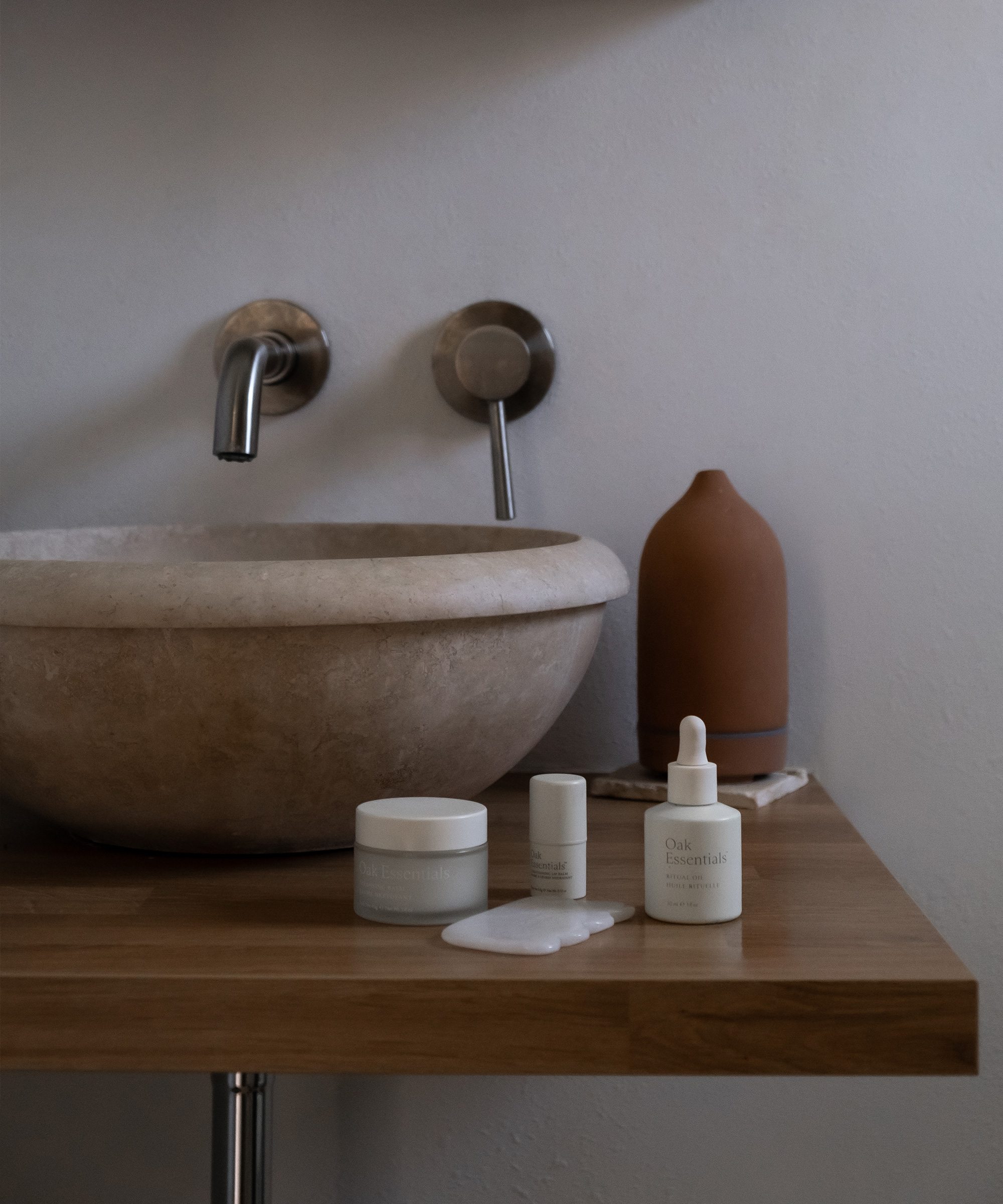 Minimalist bathroom showcases a stone sink, wall-mounted faucets, and skincare products on a wooden counter—perfect inspiration for your next room renovation. A serum bottle with some liquid spilled sits beside a terracotta vase.