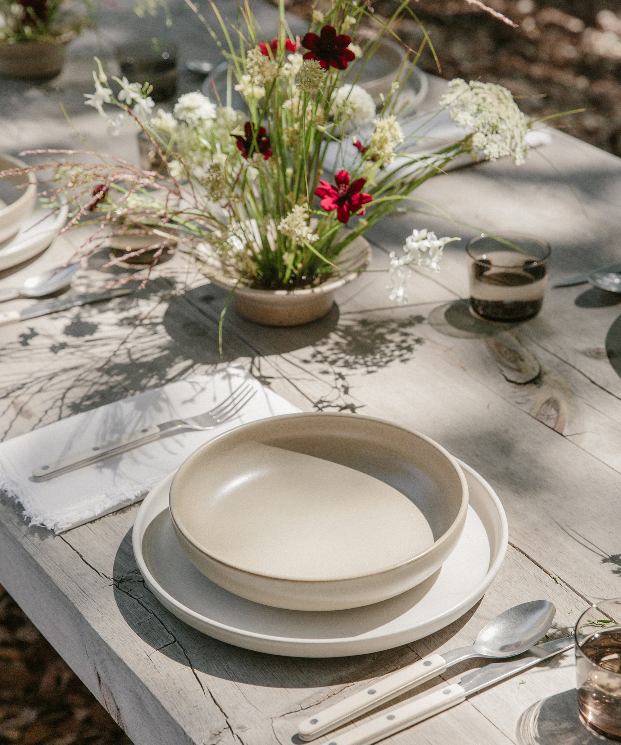 A rustic outdoor table setting with ceramic plates and bowls, white napkins, silver cutlery, and glasses. A natural flower arrangement with red and white blooms sits in the center—perfect inspiration for your next dinner party menu ideas.