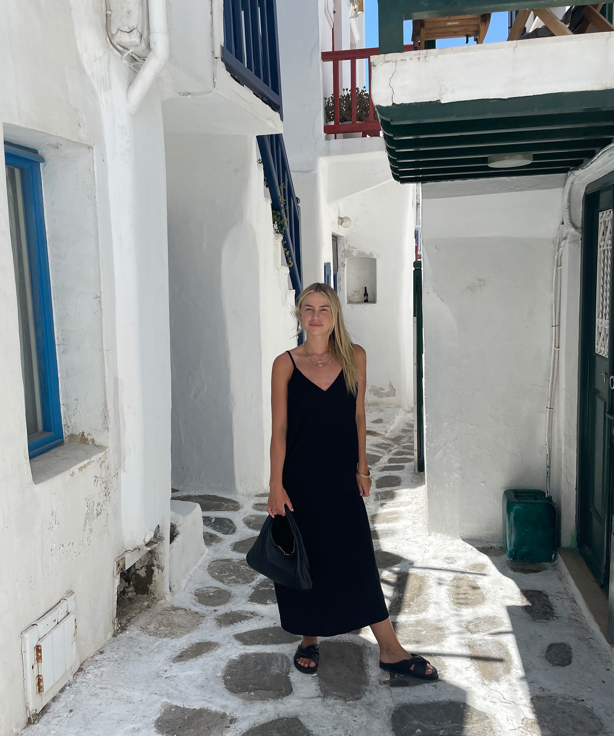 A woman in a black dress stands on a sunlit, narrow stone alleyway with white walls and blue window frames—a scene straight from a getaway guide to the Mediterranean. She holds a black bag and smiles softly at the camera.