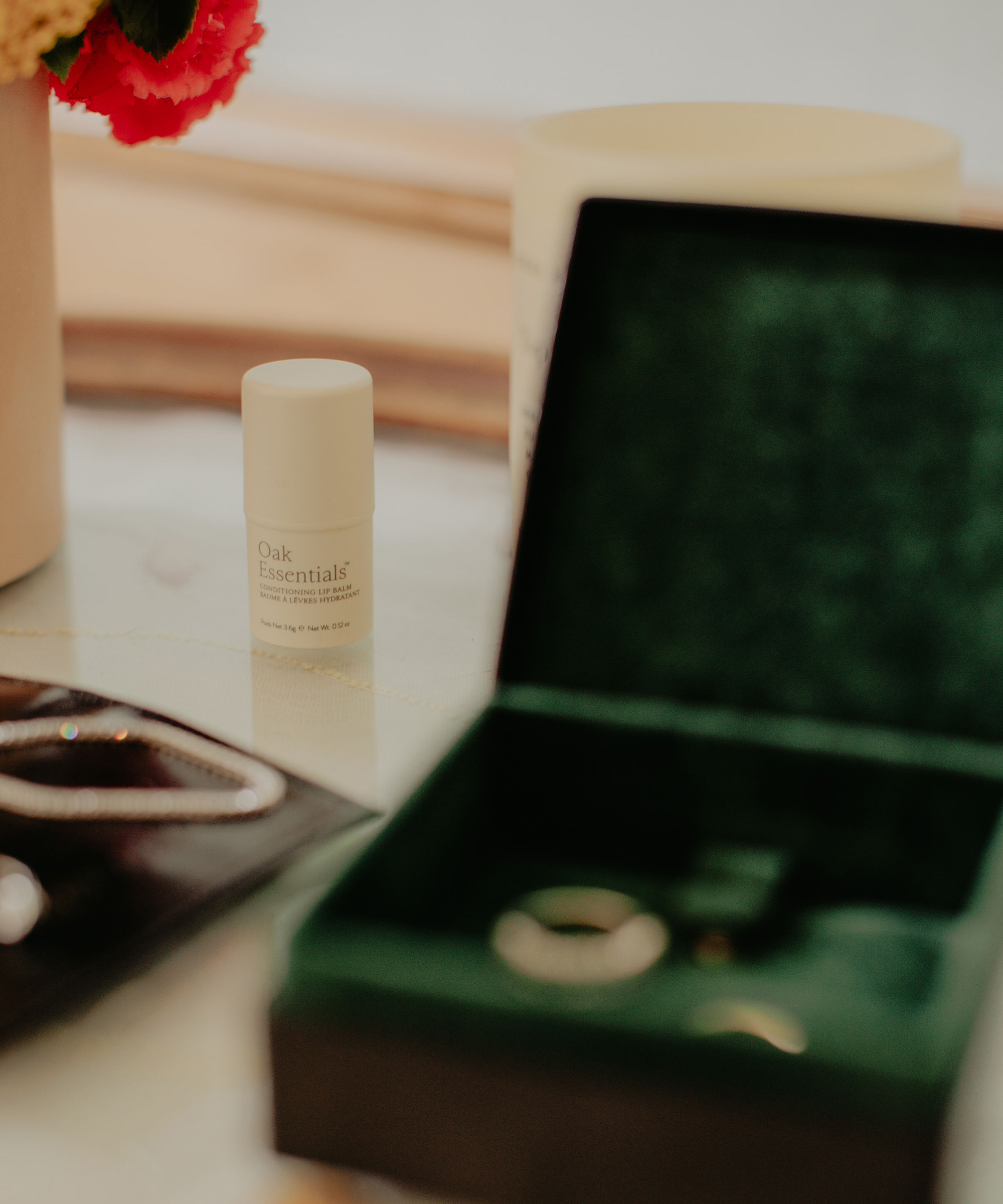 A small container labeled “Oak Essentials” sits on a table near a green velvet jewelry box with rings inside—perfect details for a destination wedding—alongside a necklace pouch and a vase with red flowers in the background.