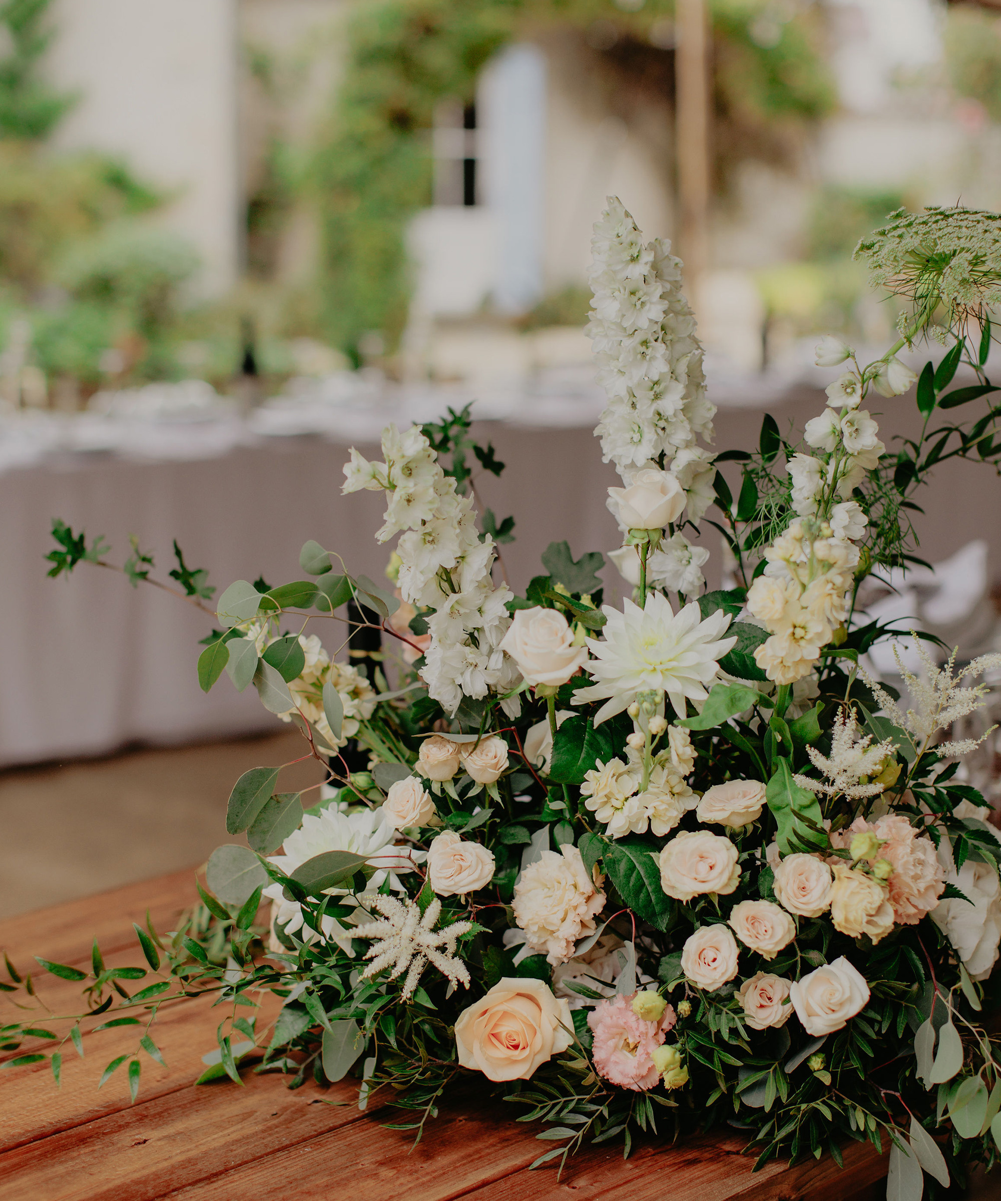 A floral arrangement of white and pale pink roses, dahlias, and greenery sits on a wooden table—perfect for a destination wedding—with a blurred outdoor background and a long table covered in a white cloth behind it.
