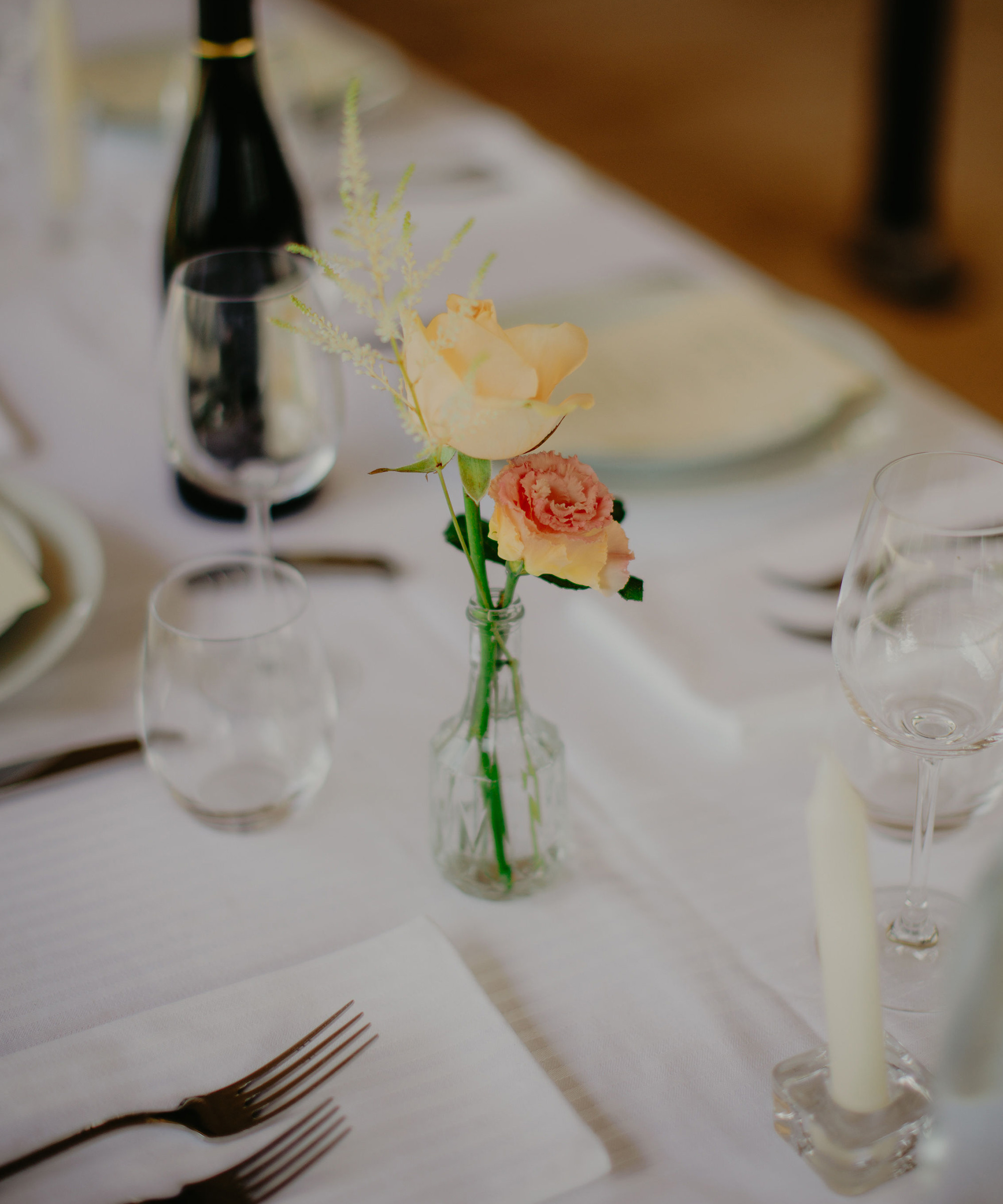 A small glass vase with pale yellow and pink flowers sits on a white tablecloth, surrounded by plates, glasses, cutlery, and a candle—perfect for an elegant destination wedding table setting.