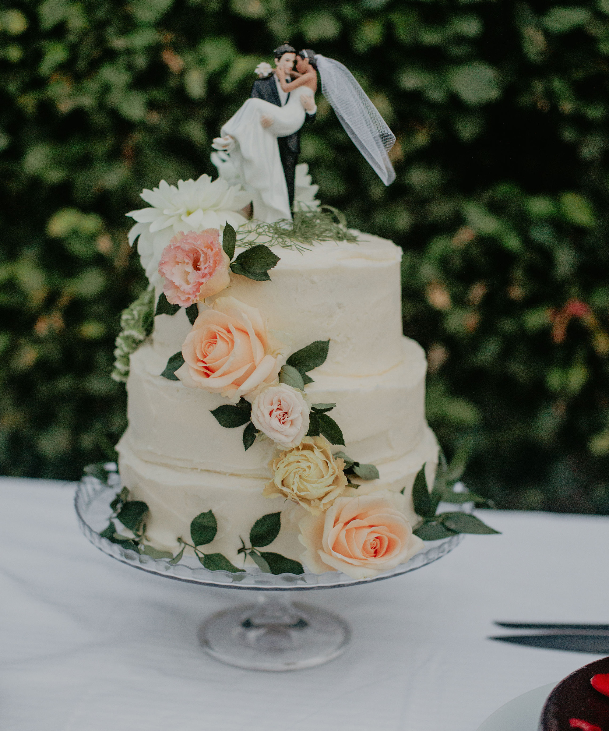 A two-tiered white wedding cake decorated with pink and peach roses, green leaves, and a bride and groom topper, perfect for a destination wedding, displayed on a glass stand with a leafy background.