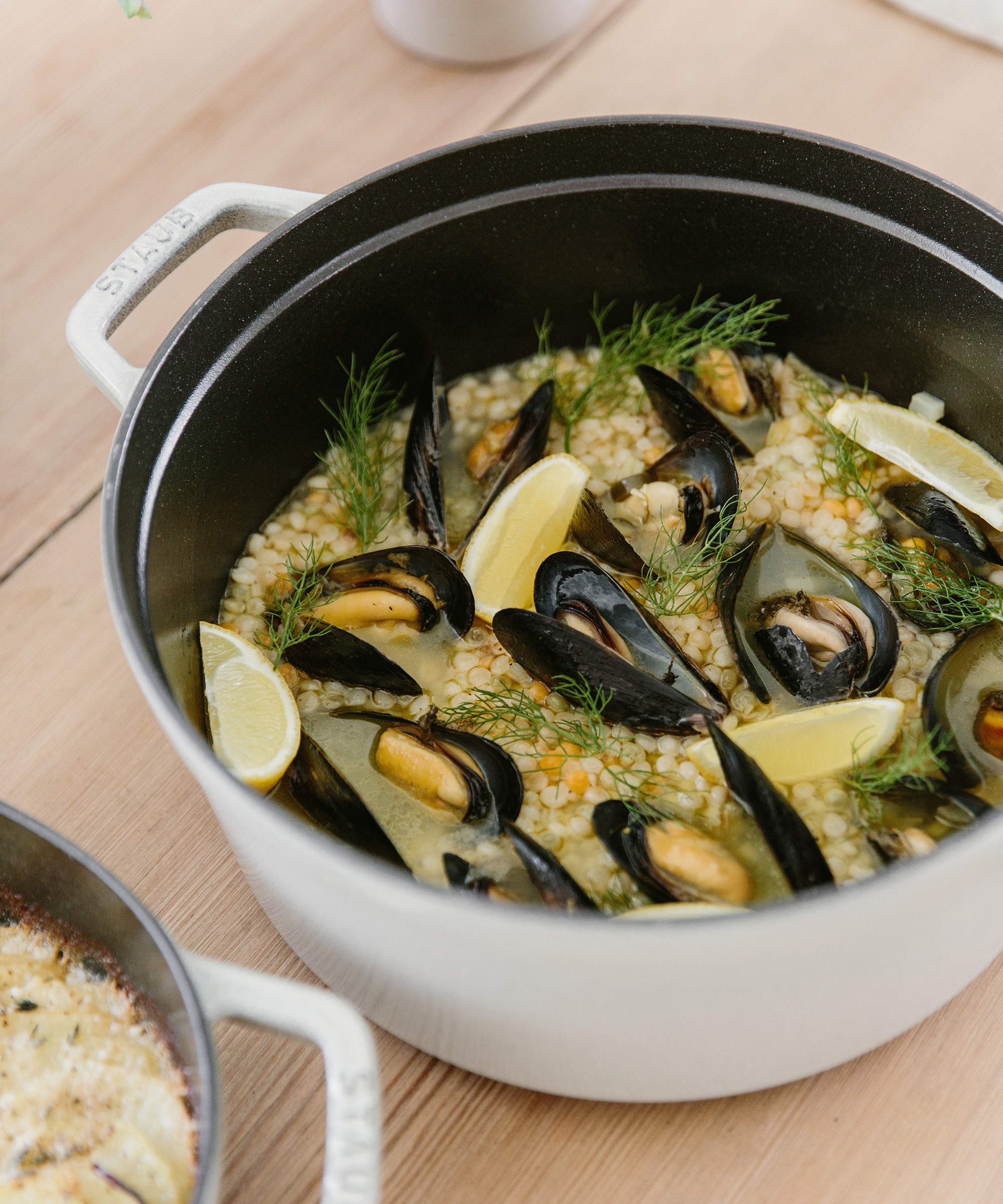 A white pot filled with cooked mussels, lemon wedges, fresh dill, and grains sits on a light wooden table—an inviting highlight for any fall menu.