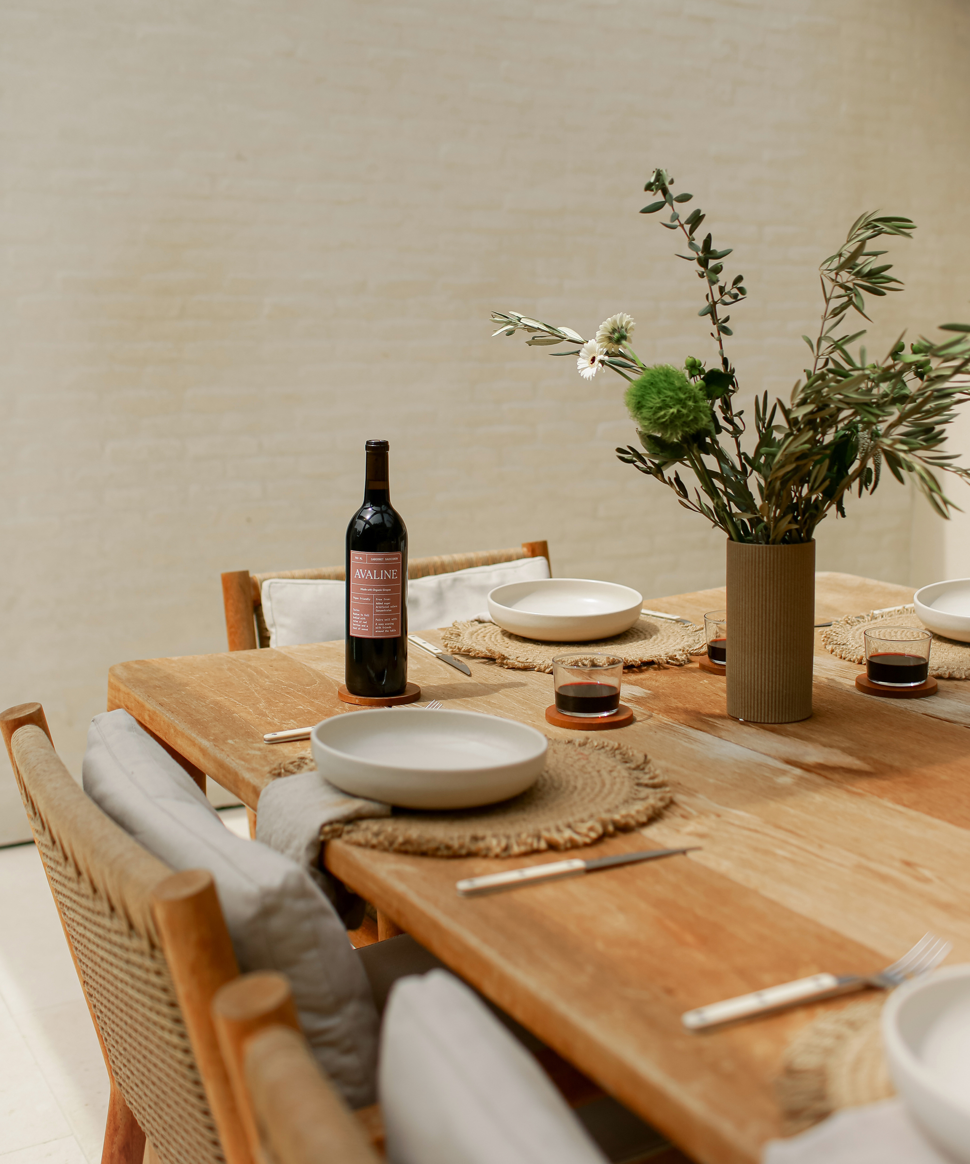 A wooden dining table set for four with woven placemats, white plates, cutlery, a vase of green and white flowers, and a bottle of Avaline red wine. The chairs have light cushions, making the setting both inviting and cozy.