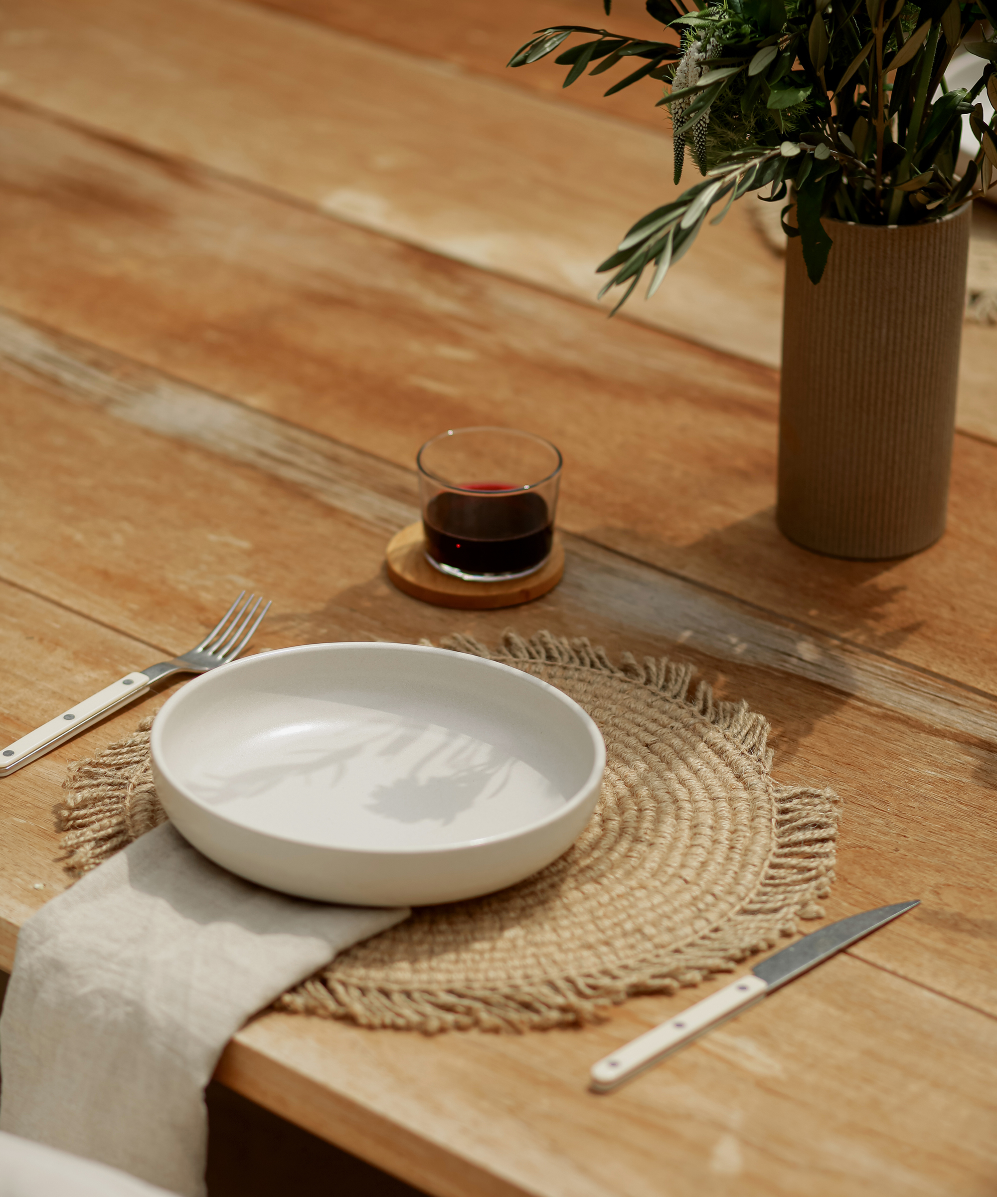 A white plate sits on a woven placemat with a fork, knife, and beige napkin on a wooden table. An Avaline red drink glows in the glass beside a vase filled with green foliage in the background.