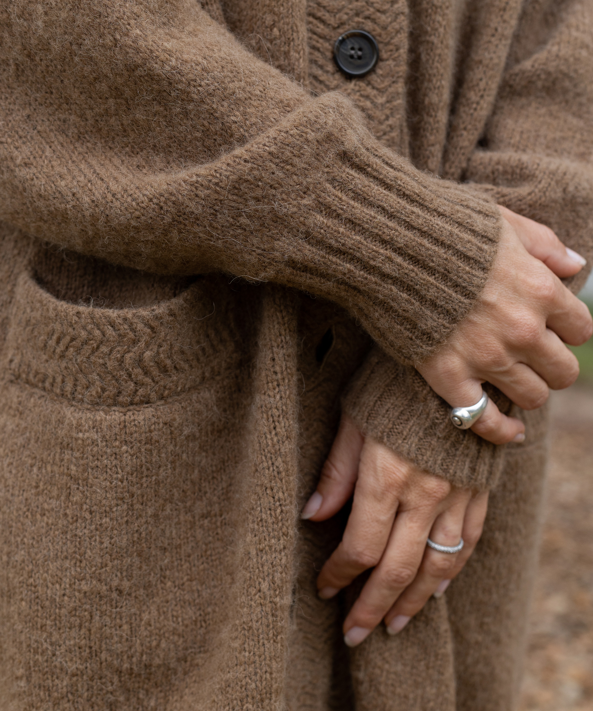 Close-up of a person’s hands, adorned with rings, gently crossed over each other. They are wearing a soft, brown knit cardigan—an essential fall uniform—with a pocket and visible buttons. The background is blurred.