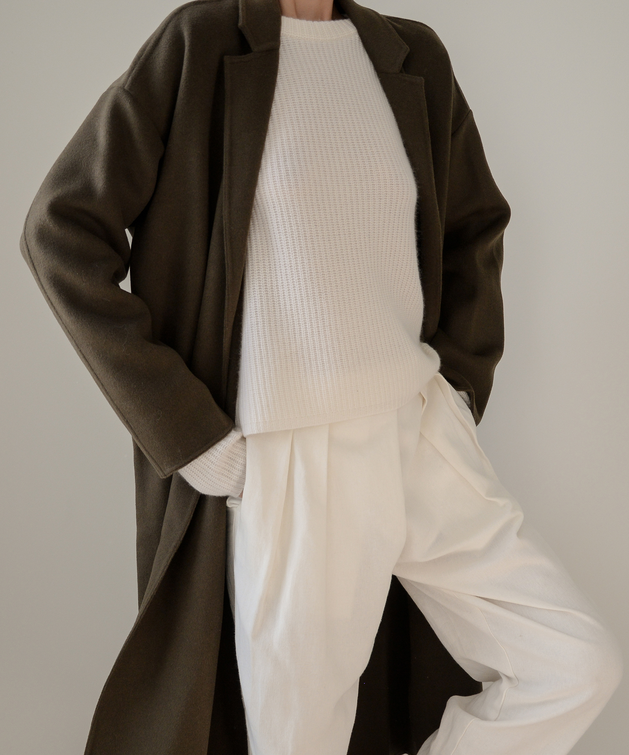 A person dressed in a long, dark coat over a cream knit sweater and high-waisted, loose white pants embodies minimalist style, standing with one hand in their pocket against a plain background.