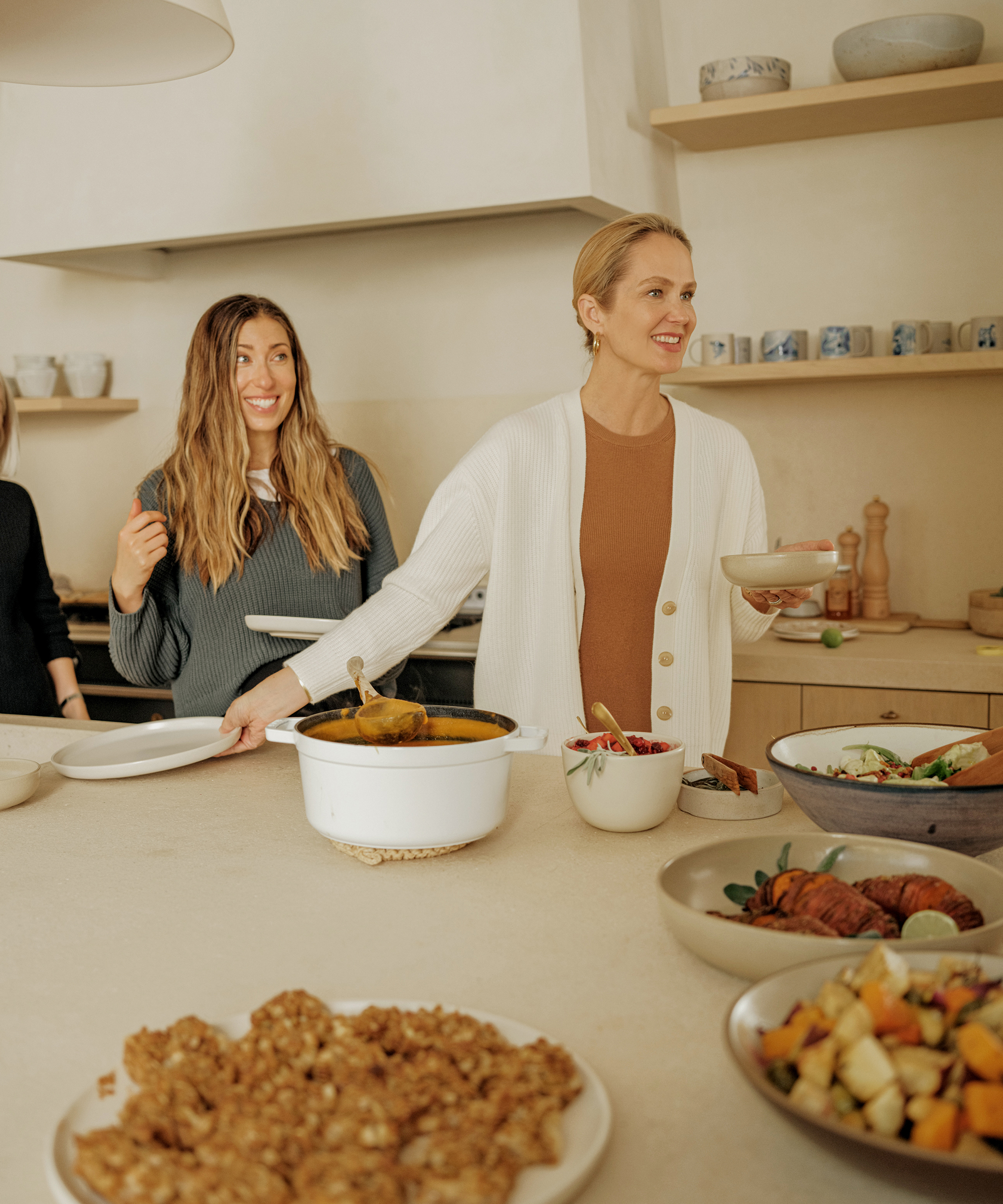 Two women stand in a kitchen serving themselves from a tempting Thanksgiving menu spread on the counter. One holds a plate and smiles, while the other, also smiling, reaches for food among various bowls and dishes.