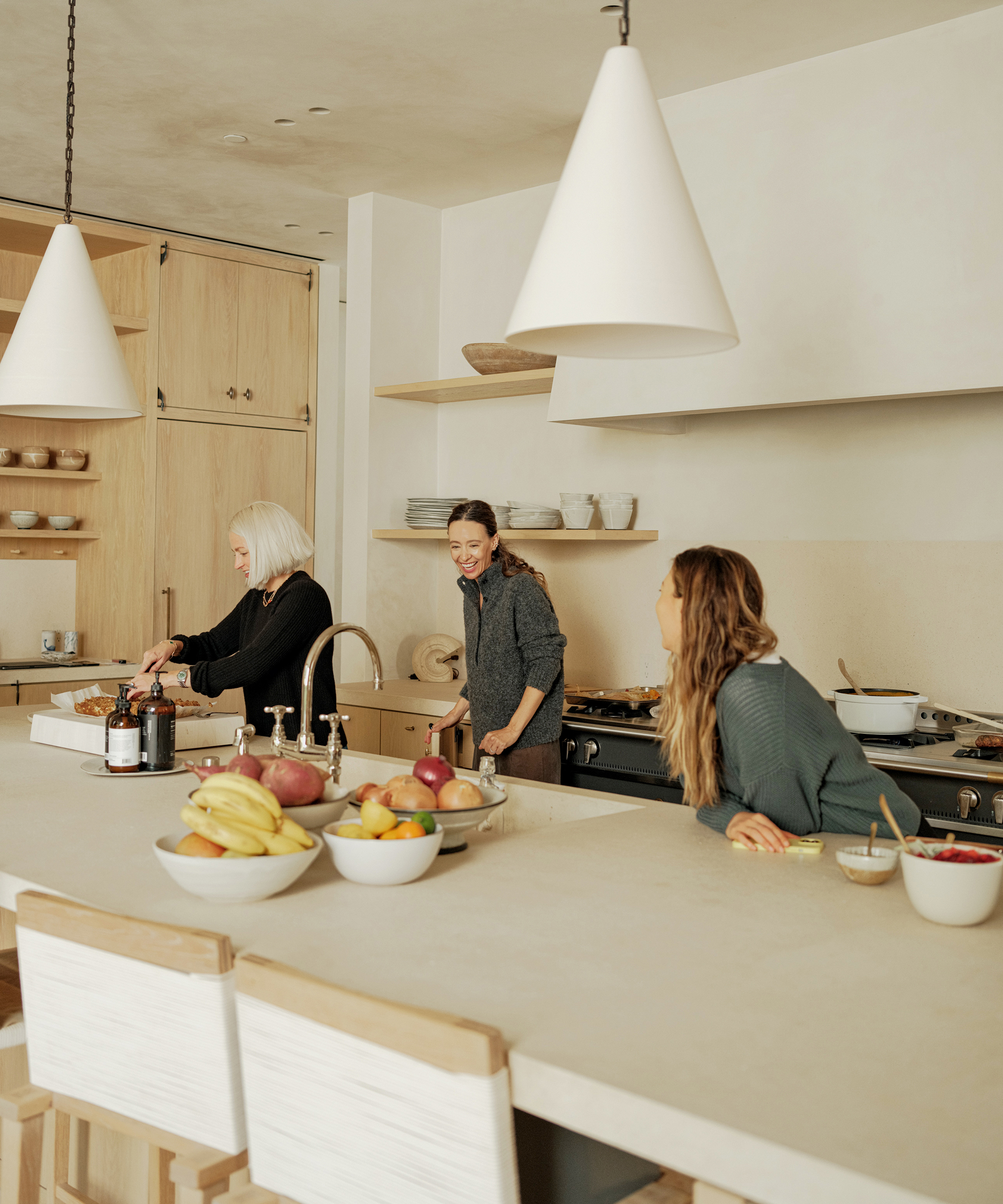 Three women enjoy cooking and talking together in a bright, modern kitchen, planning their Thanksgiving menu amid wooden cabinets, white countertops, and bowls of fruit on the island.